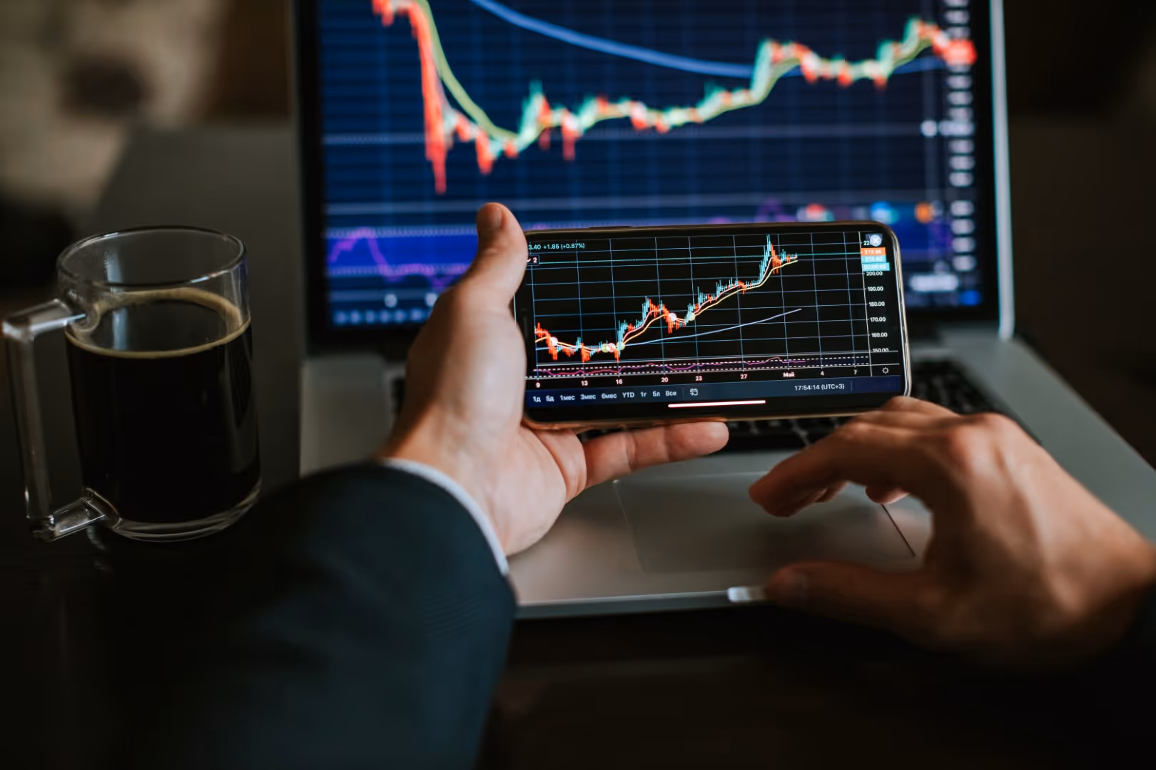 A person holding a phone and trading in front of a laptop displaying trading charts and market data.