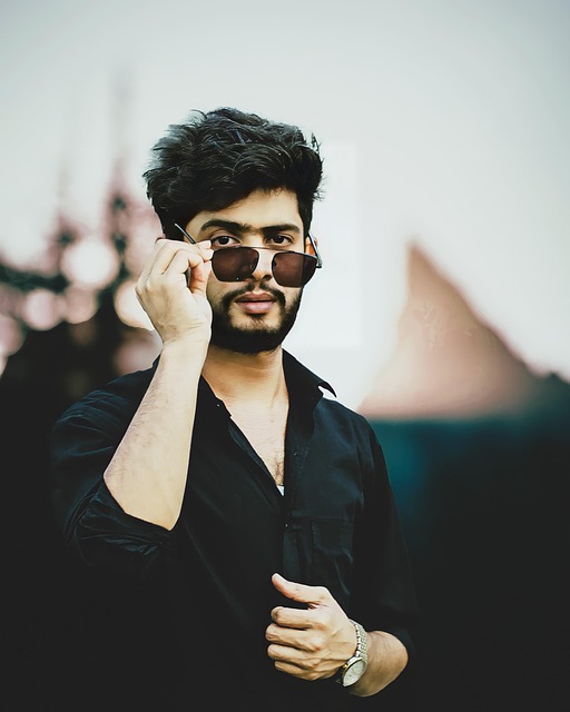 Jeune homme portant une chemise noire retirant ses lunettes de soleil avec un arrière-plan flou.