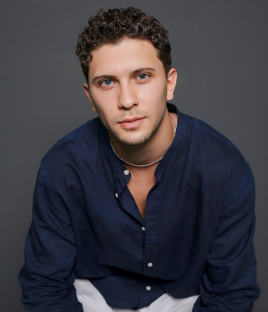 Portrait d'un homme aux cheveux bouclés, portant une chemise bleu foncé avec un collier simple, fond gris.