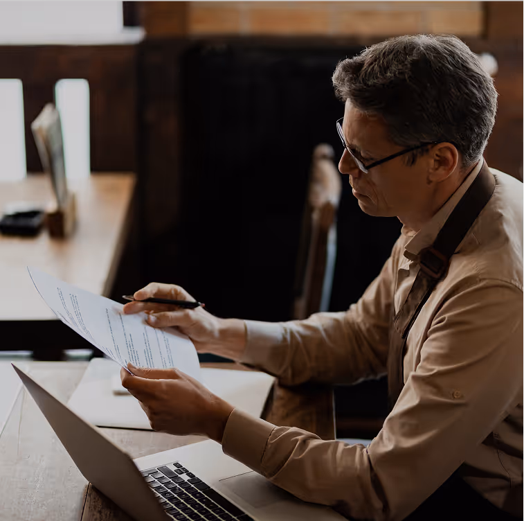 Middle-aged man wearing glasses and apron reviewing papers while sitting at a wooden desk with an open laptop.