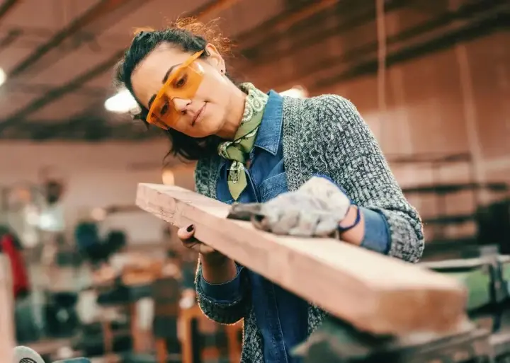 Craftsperson wearing safety glasses measures wooden plank in workshop