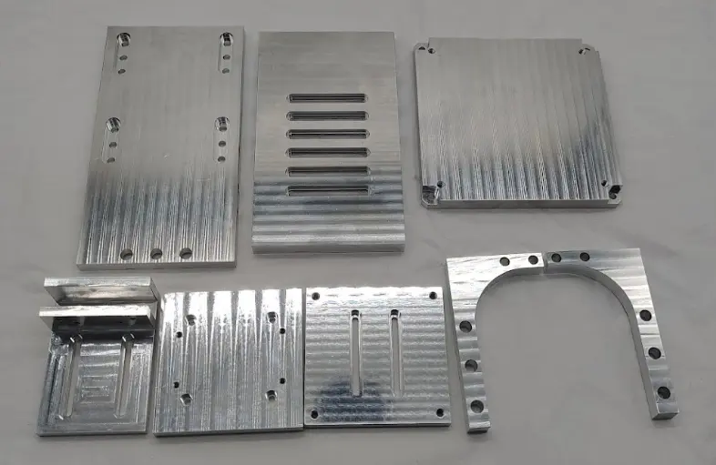 Set of CNC-machined aluminum parts arranged on a flat surface, showcasing various milling patterns, slots, and drilled holes.
