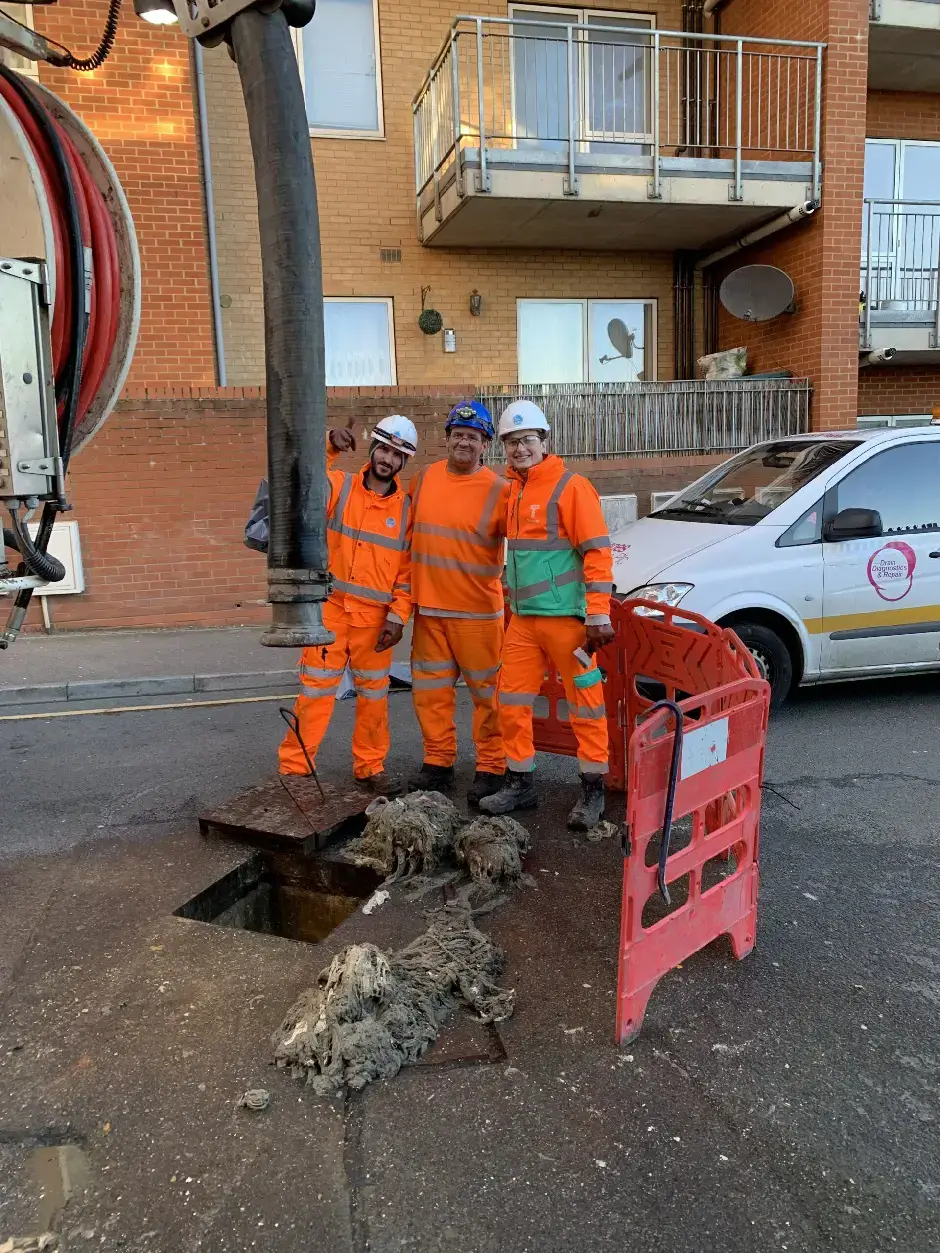 Three utility workers in orange safety gear standing near open street excavation