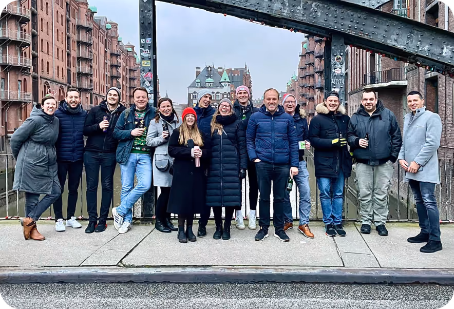 A Digimondo team standing outside in a city, smiling and posing for a photo. They are wearing various clothing styles, including jeans and footwear. The sky is visible above them, and there are buildings in the background.