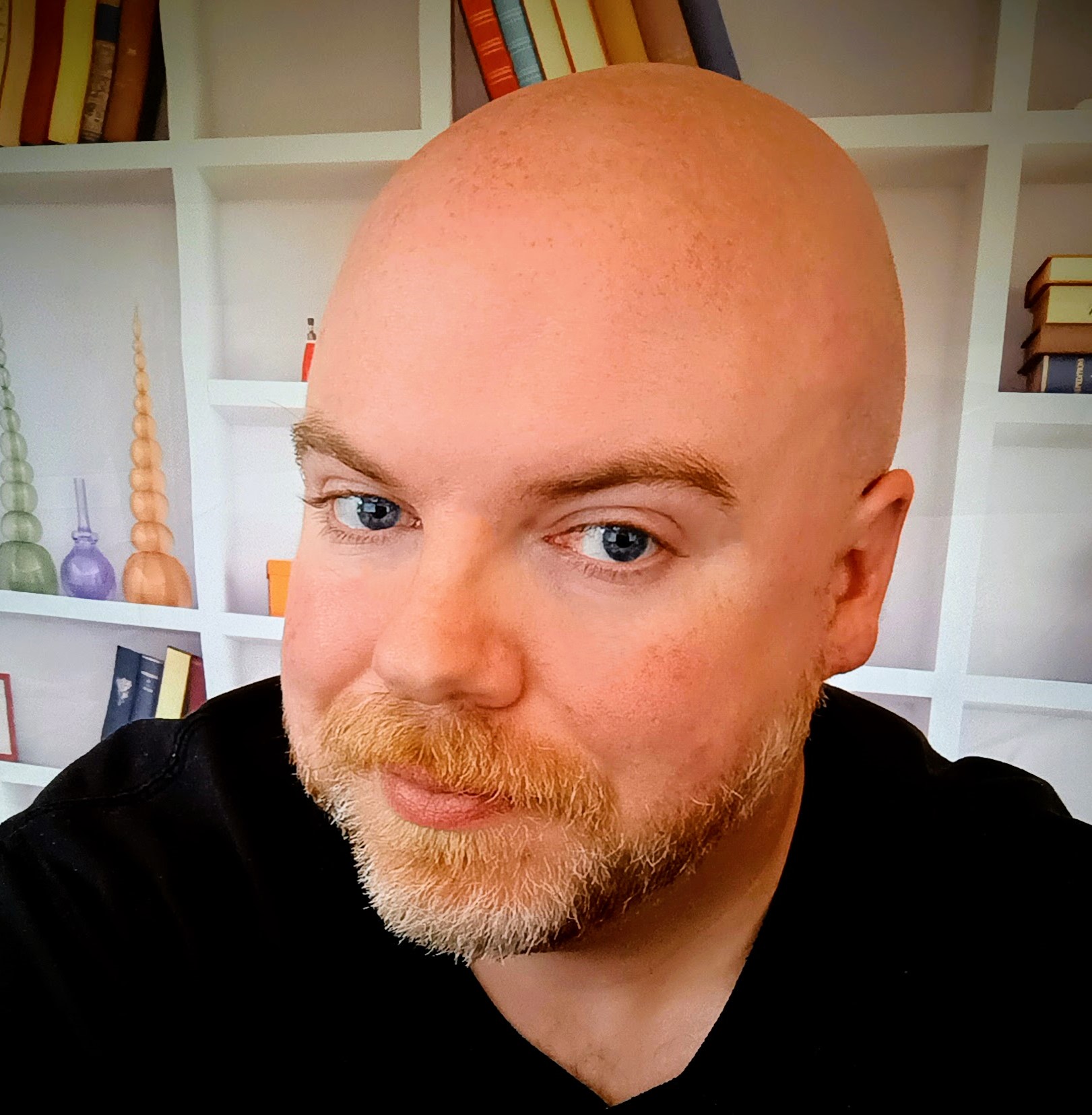 A photo of Justin Julian from below his shoulders and up.  Wearing a black shirt and standing in front of a bookcase.  He is bald and looking to the left, with a slight beard and mustache. 