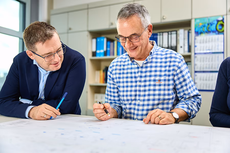 Zwei Männer stehen am Schreibtisch eines hellen Büros und diskutieren über technische Pläne.