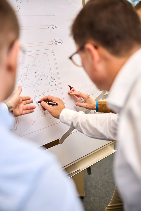Mehrere Personen analysieren eine technische Zeichnung an einem Whiteboard. Hände mit Stiften markieren und diskutieren Details auf dem Plan.