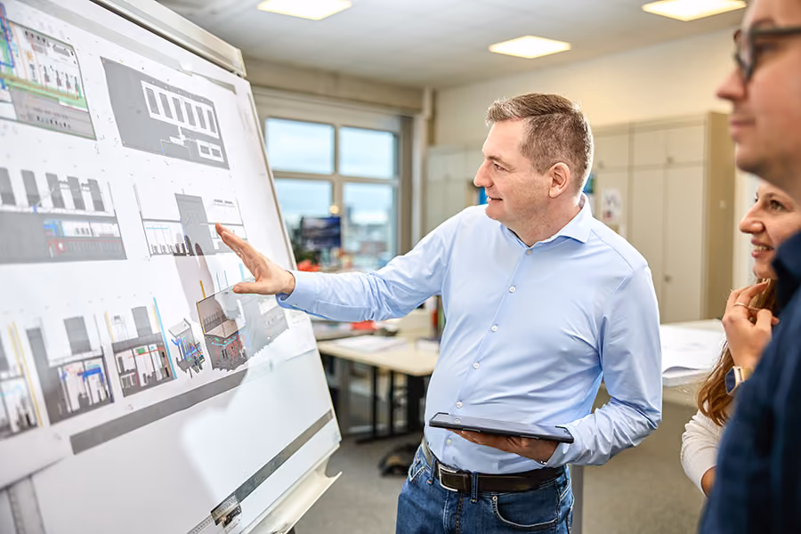 Ein Mann im Hemd steht in einem Büro, zeigt auf ein Whiteboard mit technischen Plänen und hält ein Tablet in der anderen Hand.
