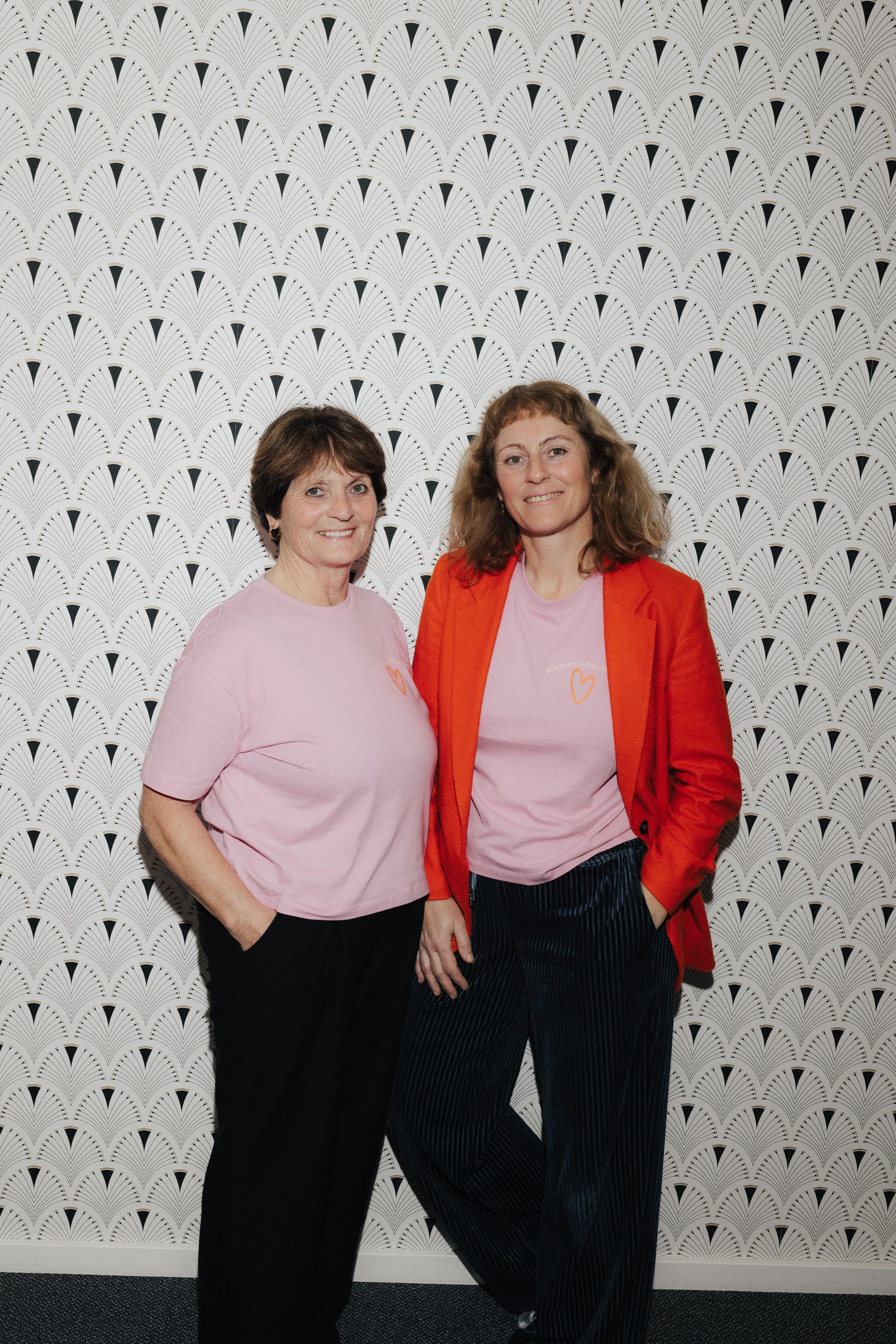 Zwei Frauen vor einer gemusterten Wand, beide tragen rosa T-Shirts; eine Frau trägt eine rote Jacke und gestreifte Hosen.