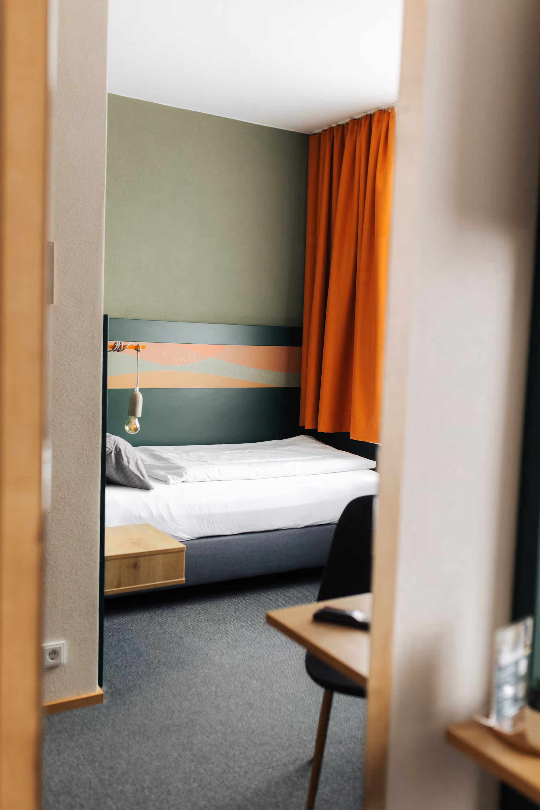 Blick durch eine offene Tür auf ein Zimmer mit Einzelbett, grünem Kopfteil, orangen Vorhängen und Schreibtisch mit Stuhl.
