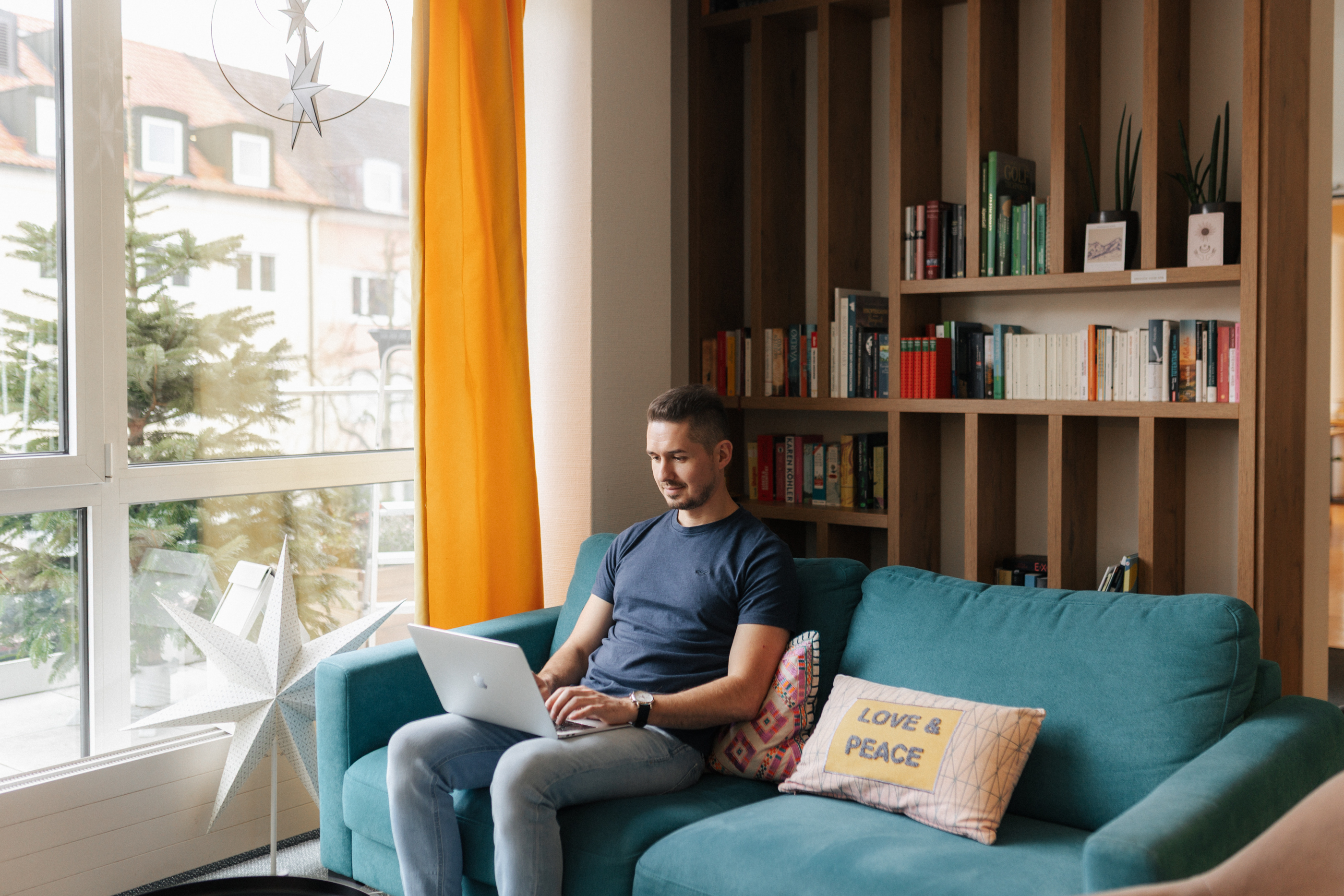Mann sitzt auf blauem Sofa mit Laptop in einem hellen Raum mit Bücherregal und gelben Vorhängen.