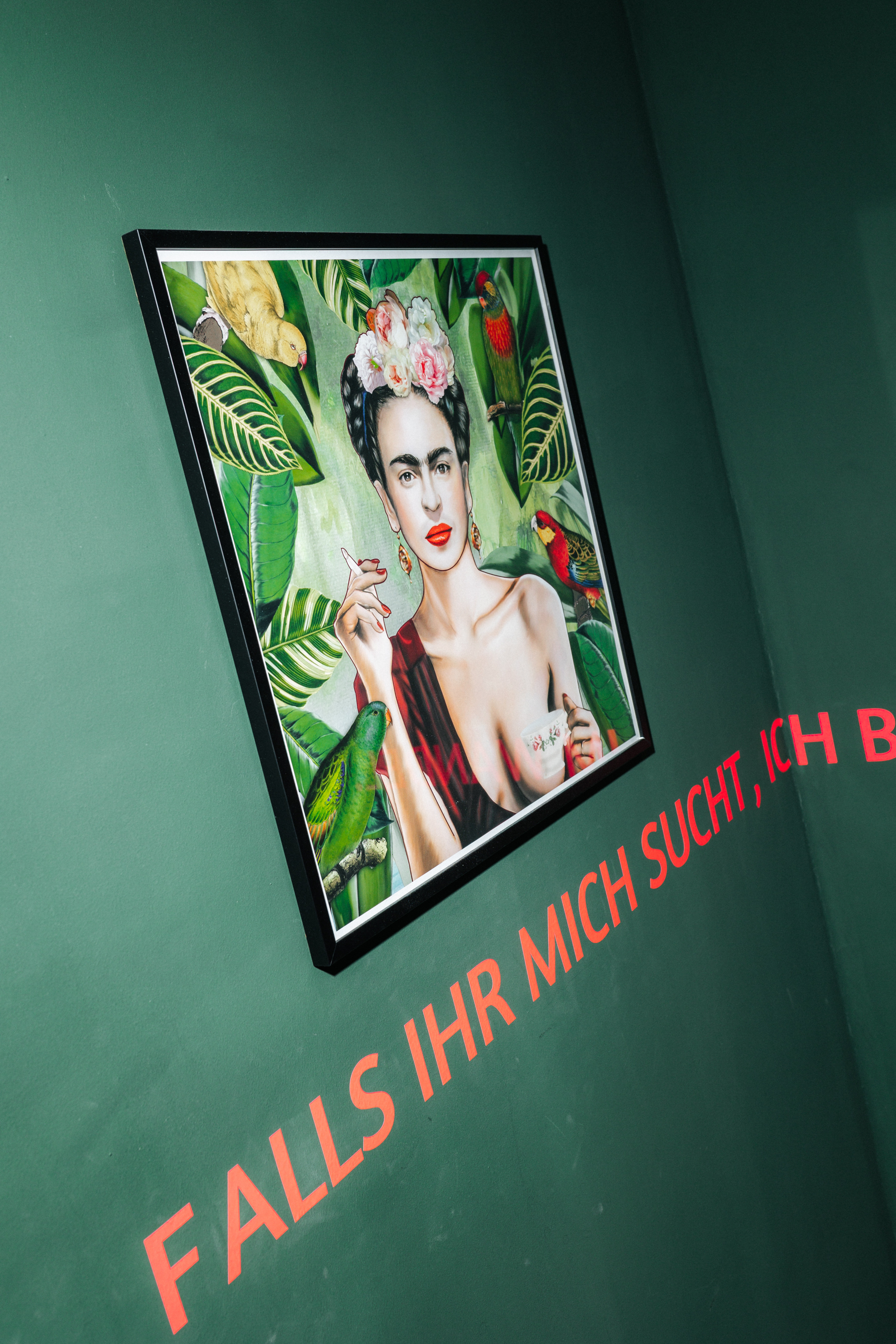 Gemälde einer Frau mit Blumen im Haar, die eine Teetasse hält und von Papageien umgeben ist, an einer grünen Wand mit rotem Schriftzug.