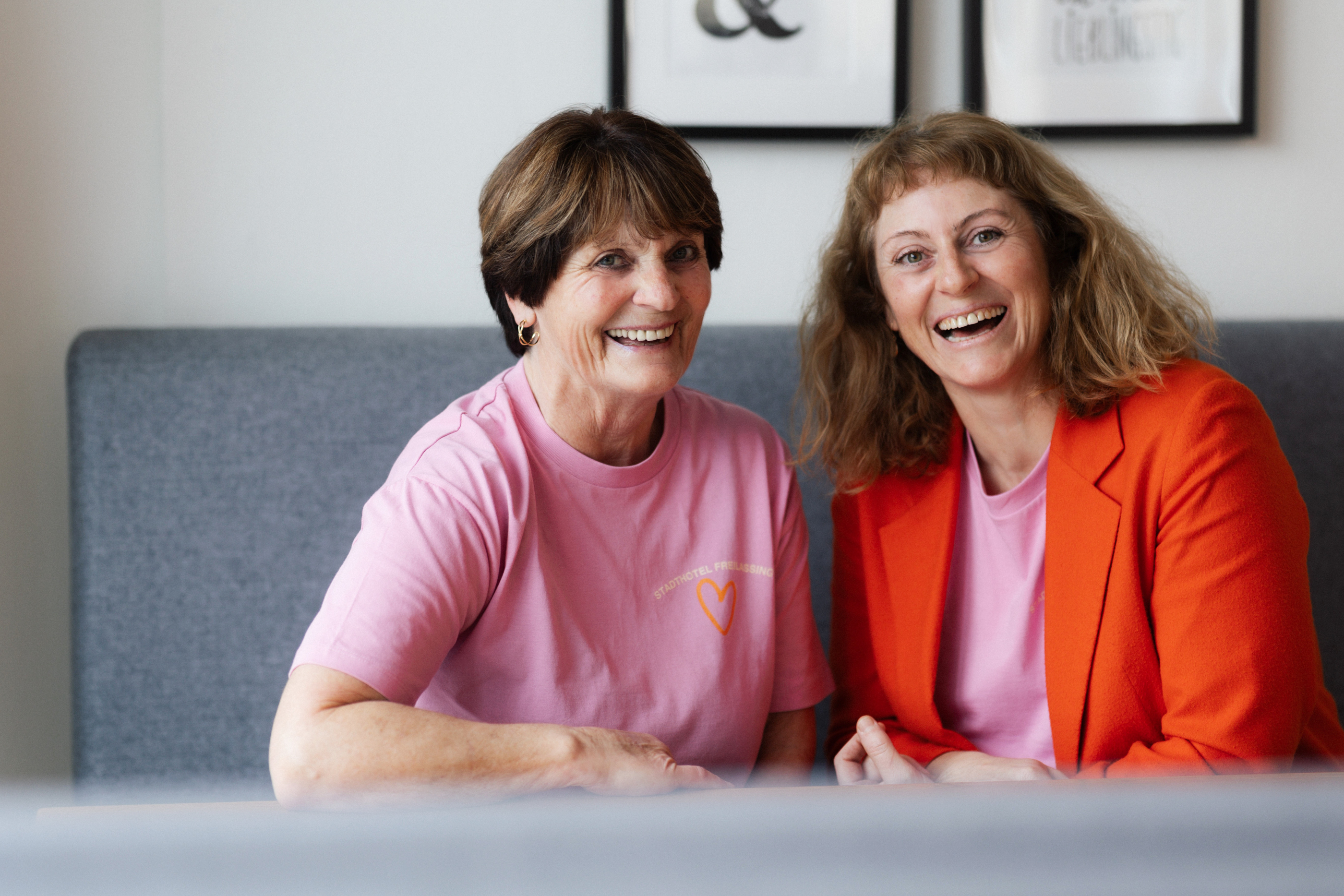 Zwei lächelnde Frauen sitzen nebeneinander auf einer grauen Bank, eine trägt ein pinkes T-Shirt, die andere ein pinkes T-Shirt mit rotem Blazer.