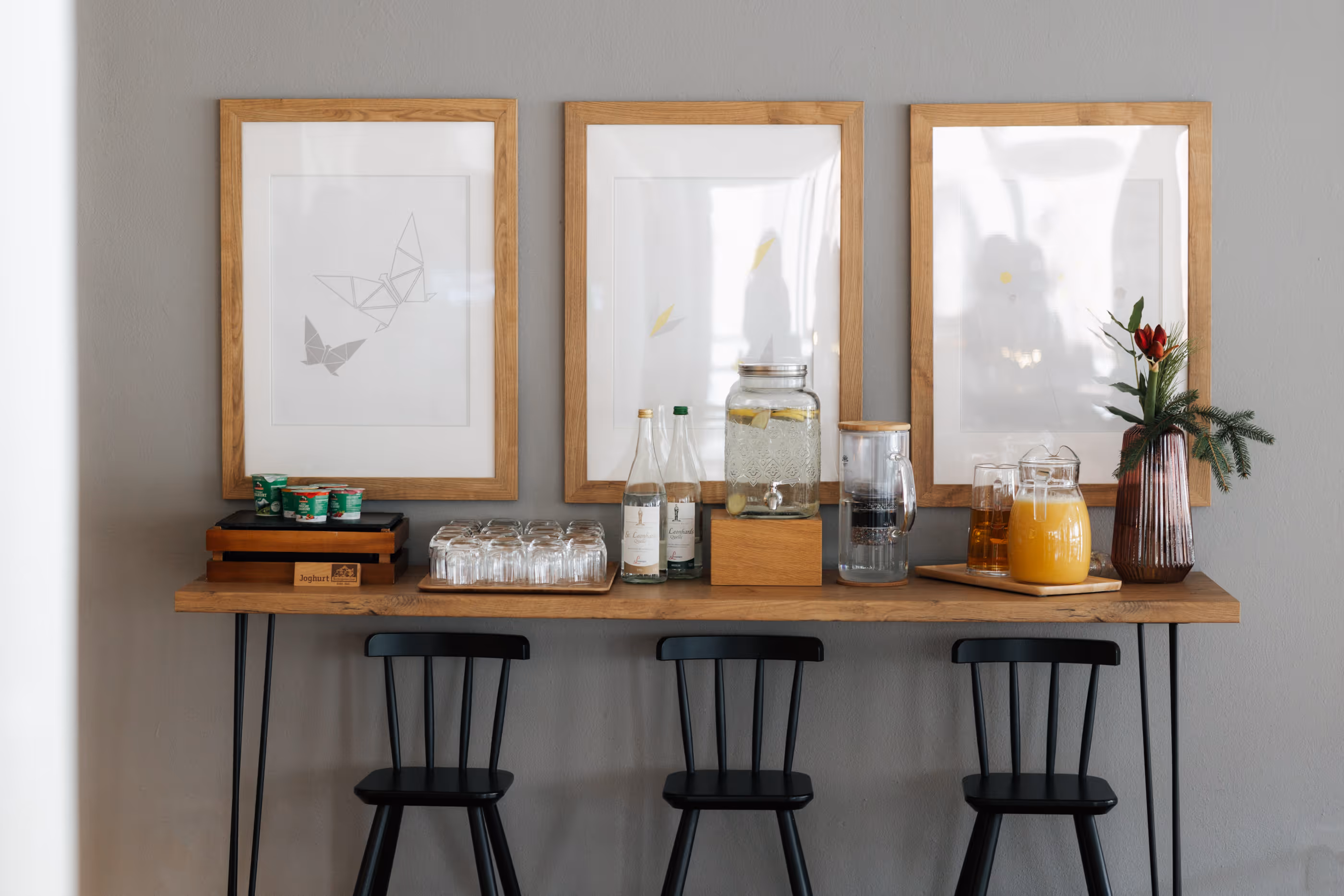 Dreierkombination aus schwarzer Barhocker, Holztheke mit Getränken und Gläsern, drei gerahmte Bilder an grauer Wand.