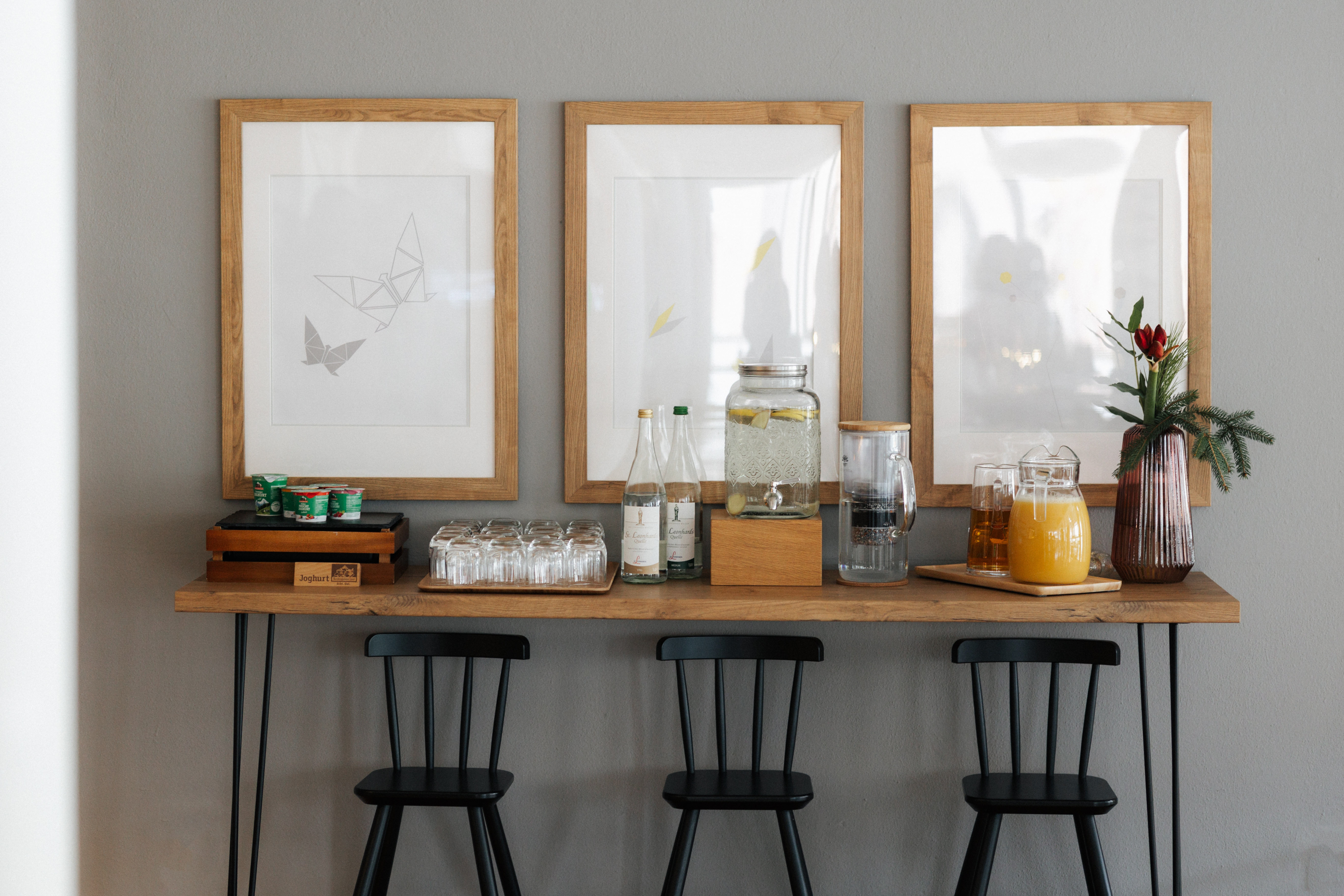 Dreierkombination aus schwarzer Barhocker, Holztheke mit Getränken und Gläsern, drei gerahmte Bilder an grauer Wand.