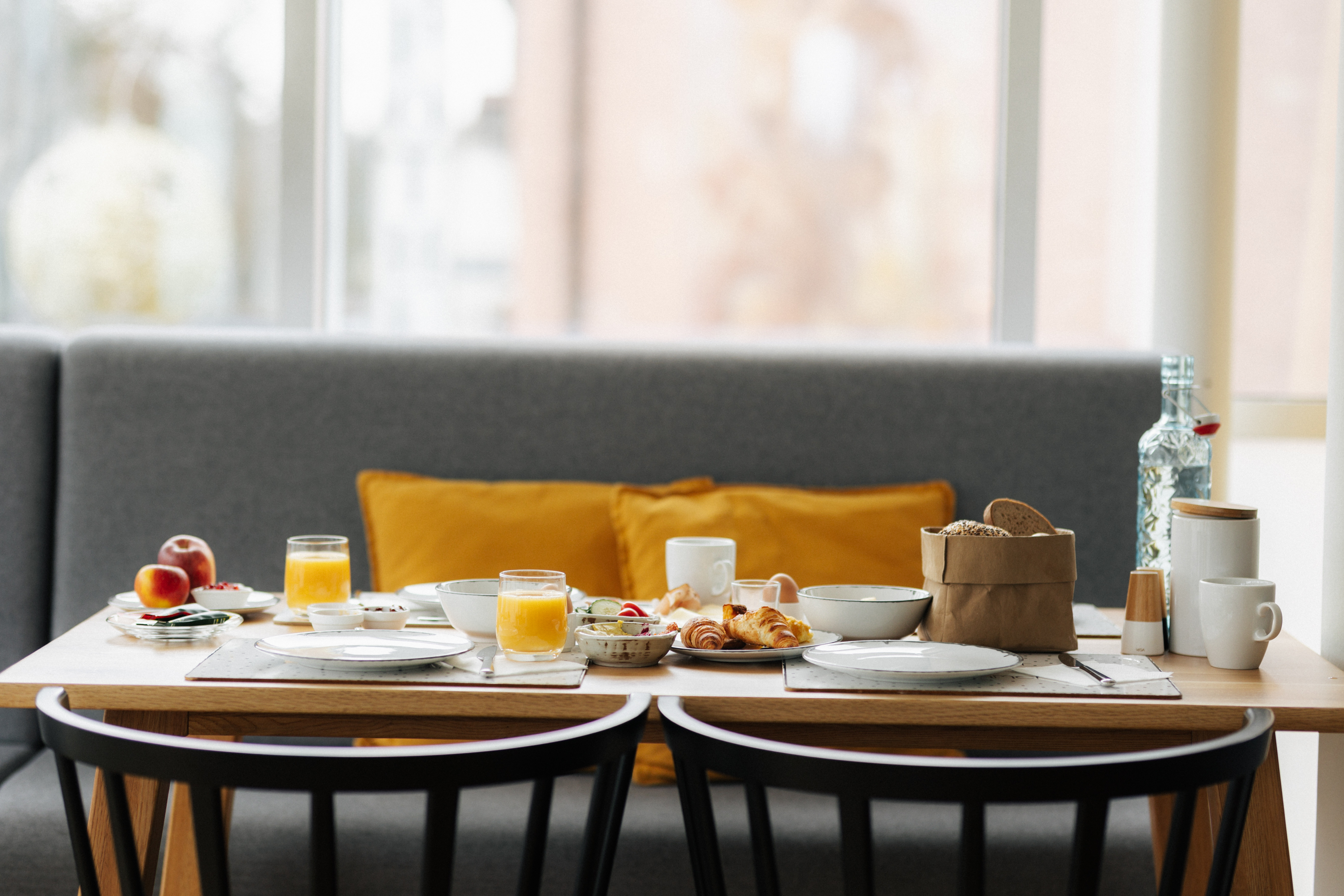 Frühstückstisch mit Orangensaft, Croissants, Brot, Äpfeln und Kaffeetassen vor grauer Sitzbank mit orangefarbenen Kissen.