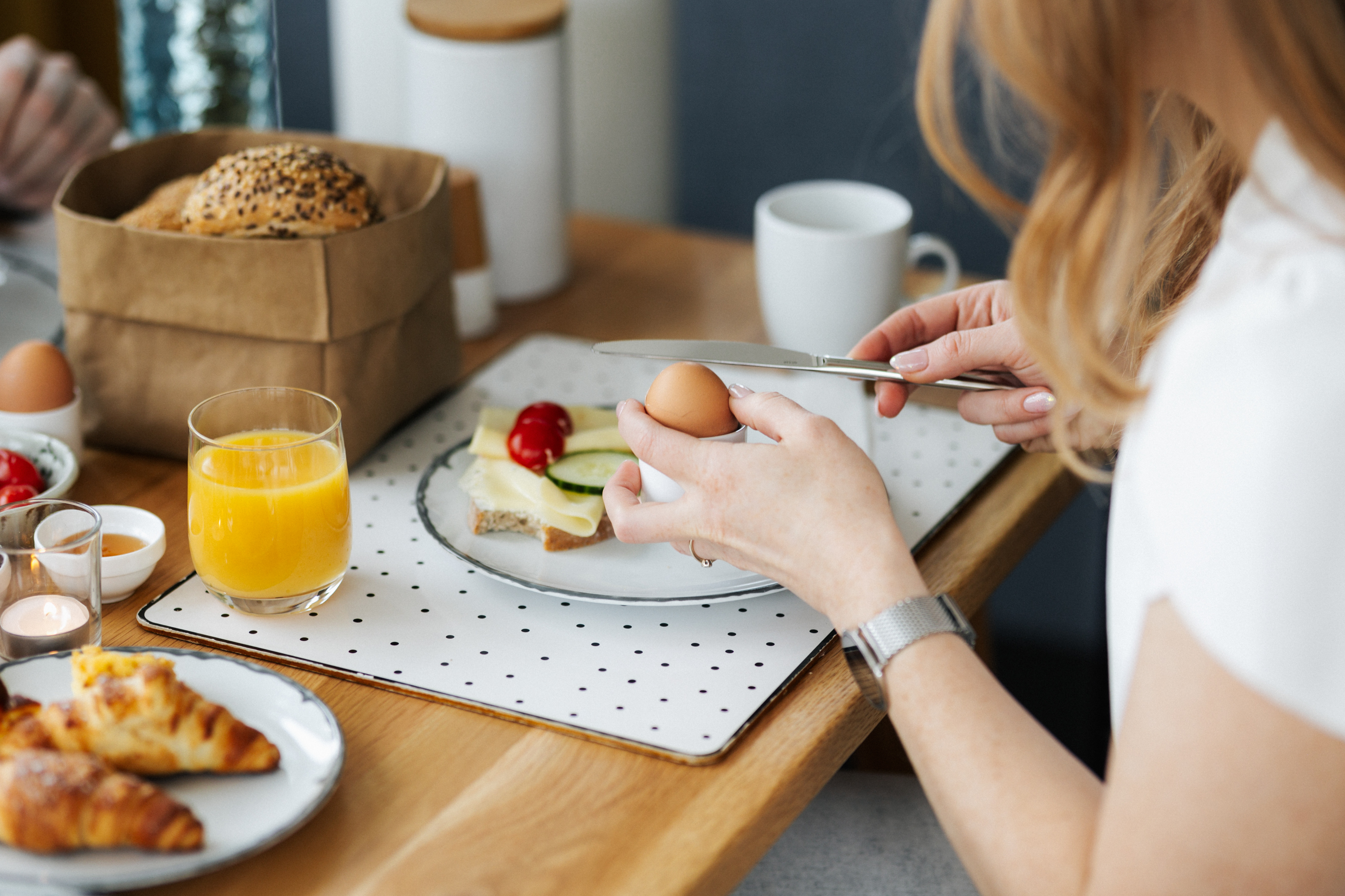 Frau schält gekochtes Ei beim Frühstück mit Croissants, Orangensaft und Käsebrot.