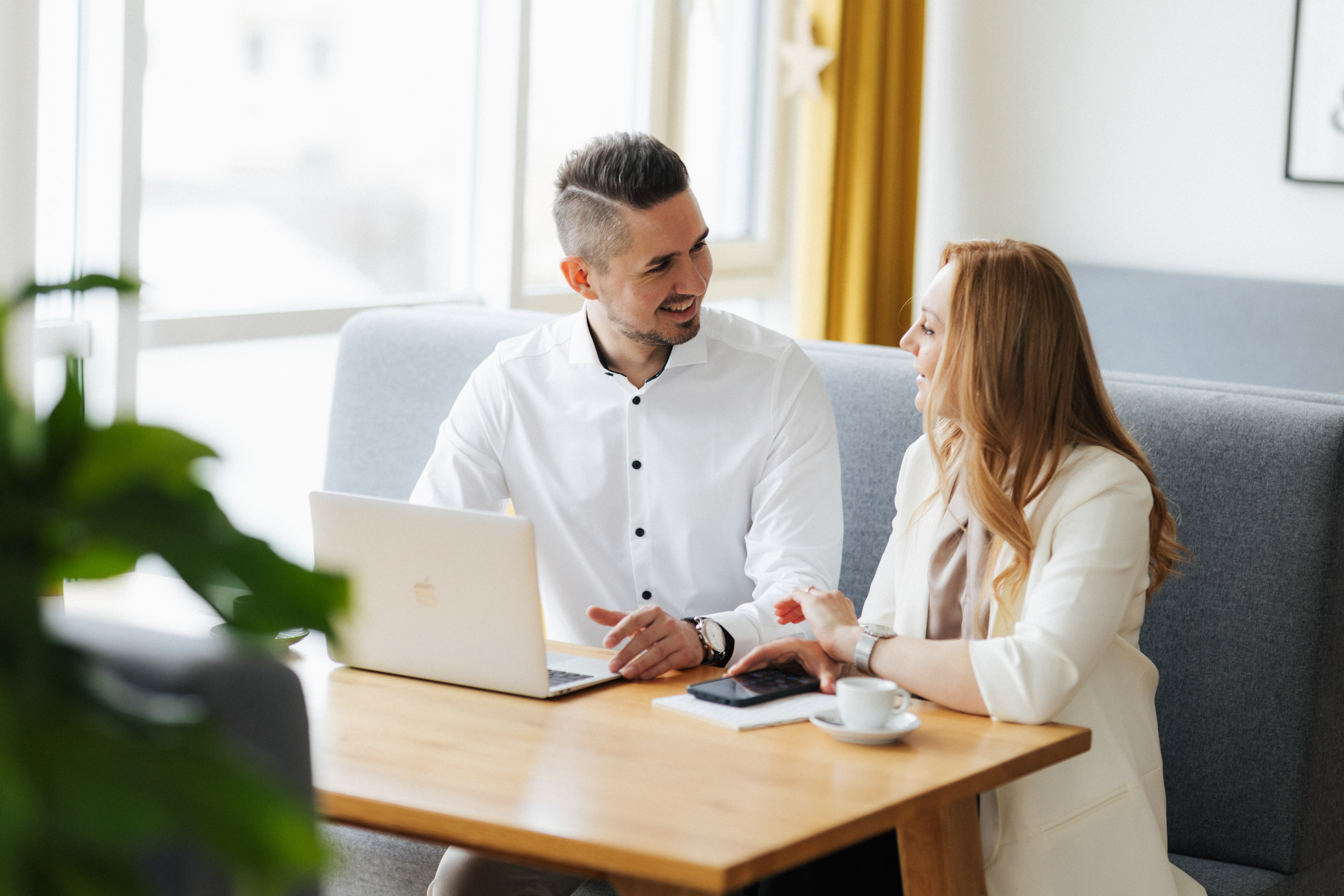 Ein Mann und eine Frau unterhalten sich lächelnd an einem Tisch mit Laptop und Kaffeetasse in einem hellen Büro.