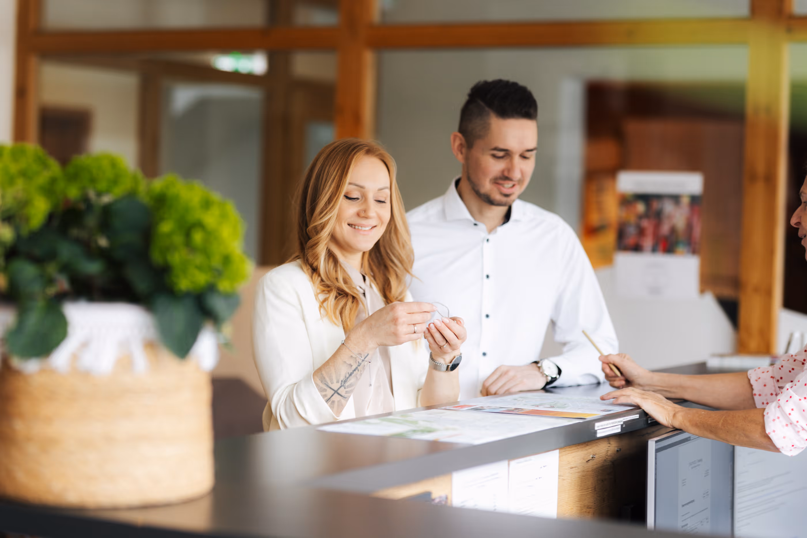 Ein Paar steht am Empfang und wird von einer Mitarbeiterin beraten, die auf einen Prospekt zeigt.