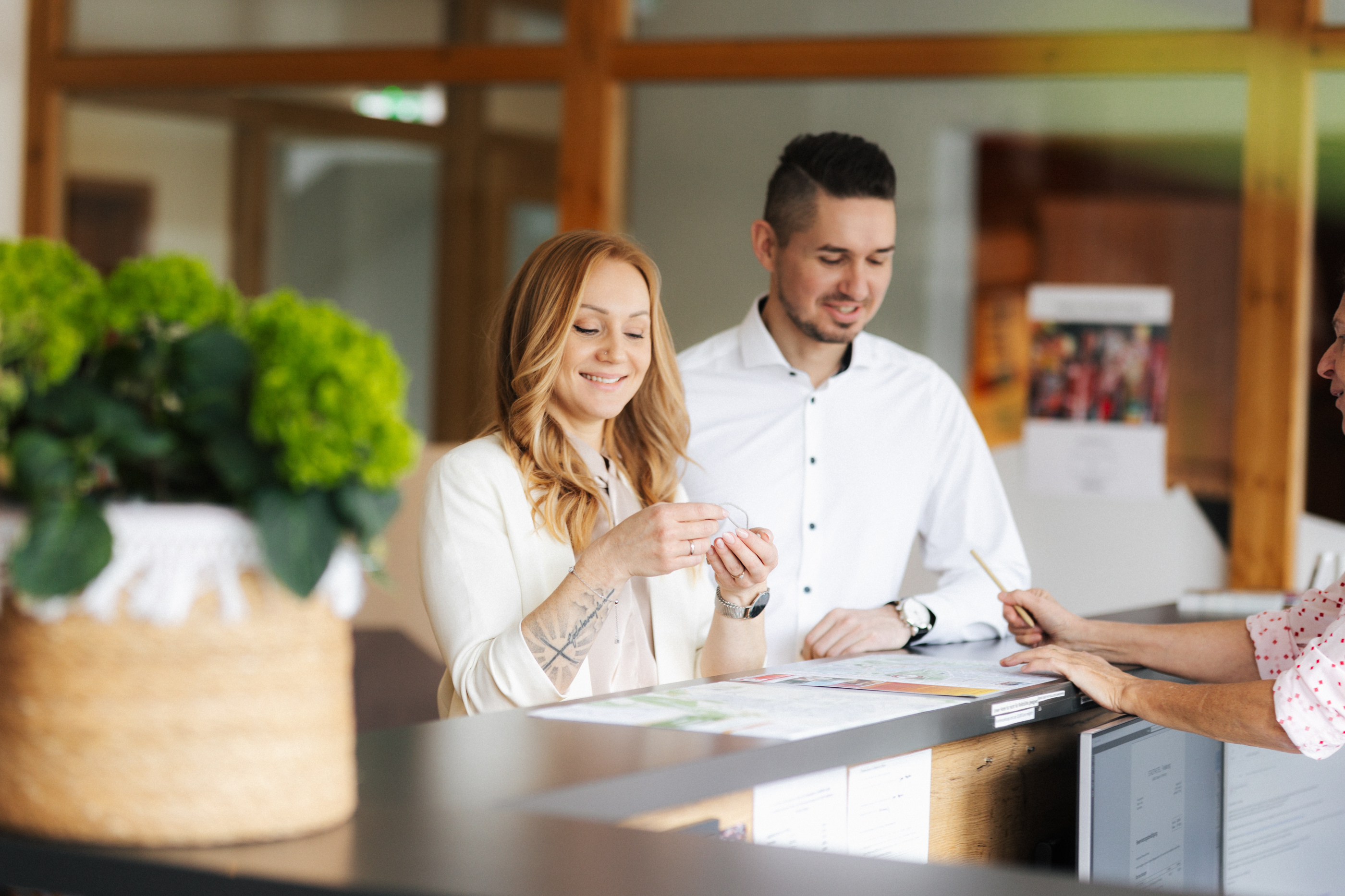 Ein Paar steht am Empfang und wird von einer Mitarbeiterin beraten, die auf einen Prospekt zeigt.