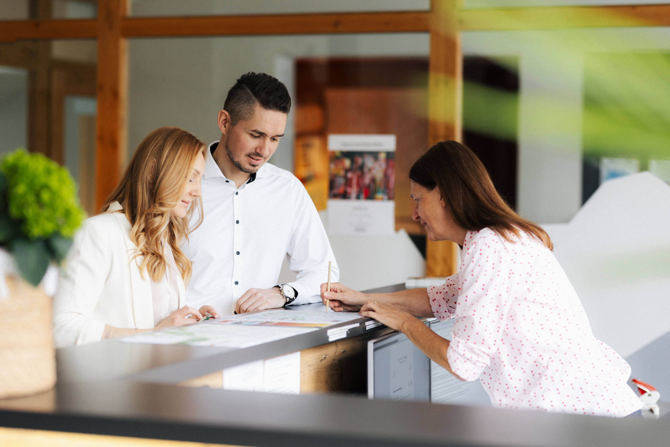 Paar wird an einer Rezeption von einer Mitarbeiterin im weißen Hemd mit roten Punkten betreut.