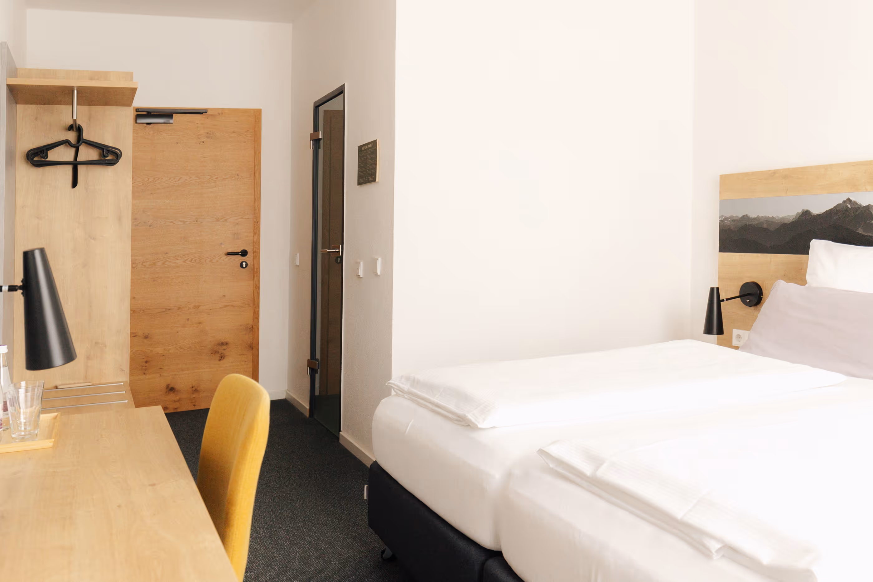 Modernes Hotelzimmer mit zwei Einzelbetten, Holzschreibtisch, gelbem Stuhl und Garderobe mit schwarzen Kleiderbügeln.