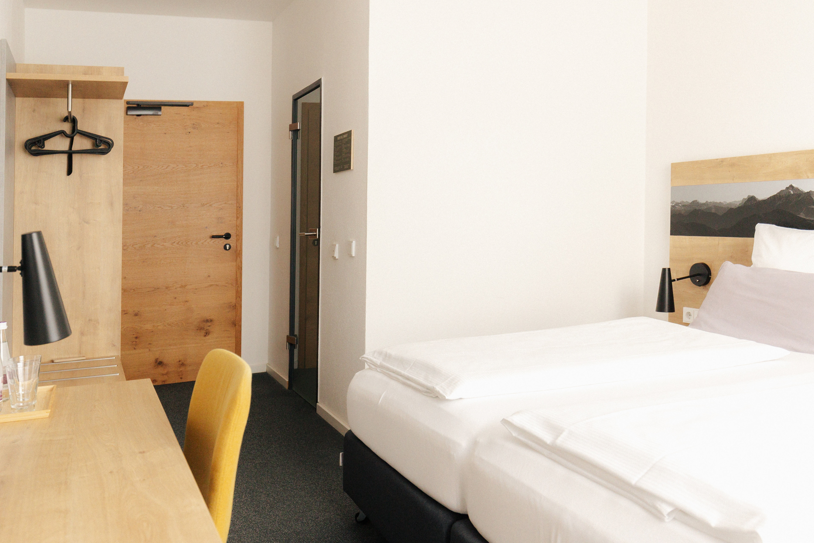 Modernes Hotelzimmer mit zwei Einzelbetten, Holzschreibtisch, gelbem Stuhl und Garderobe mit schwarzen Kleiderbügeln.