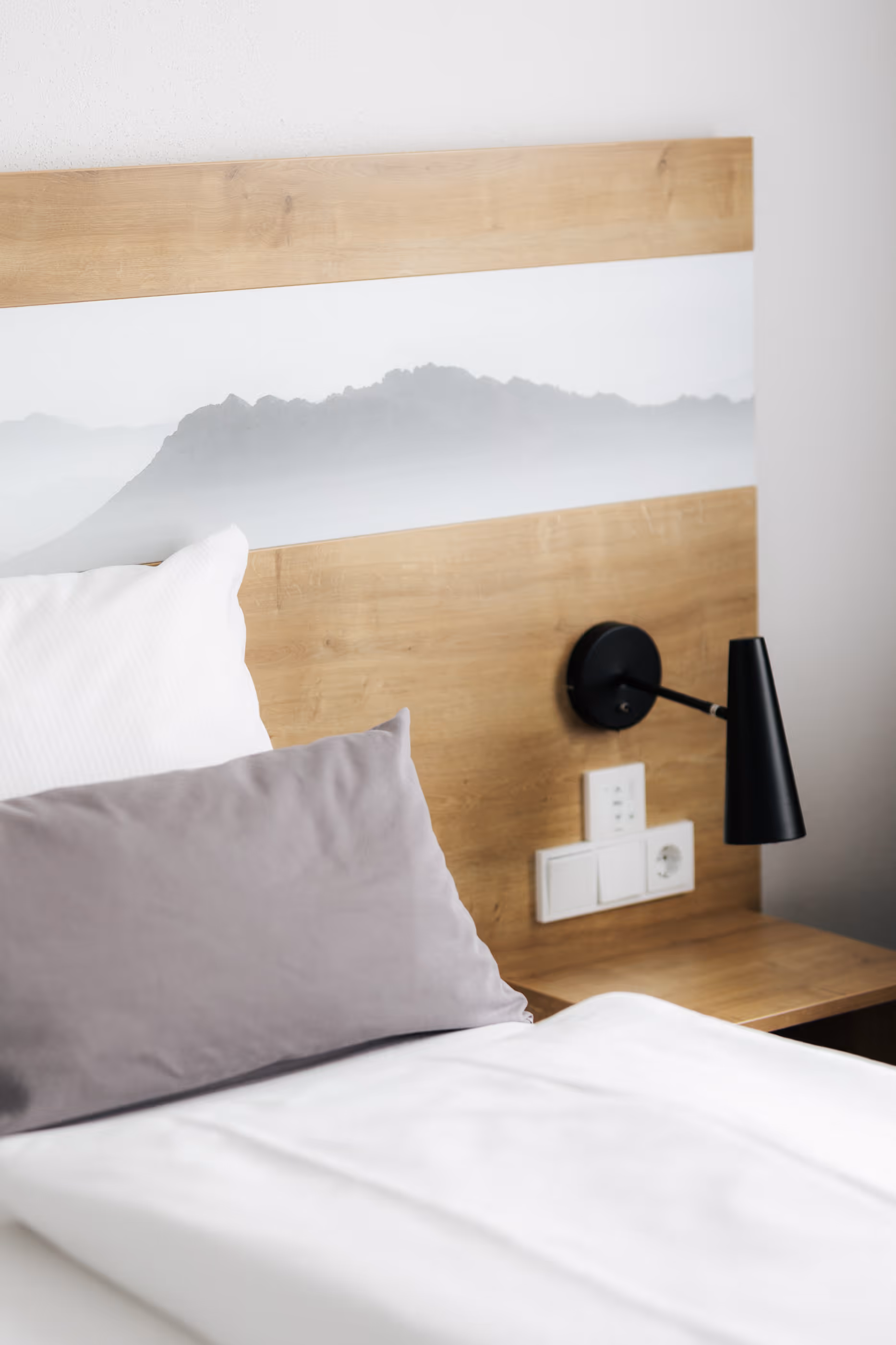 Bett mit weißen und grauen Kissen, Holz-Kopfteil mit Berglandschaft und schwarze Wandlampe.