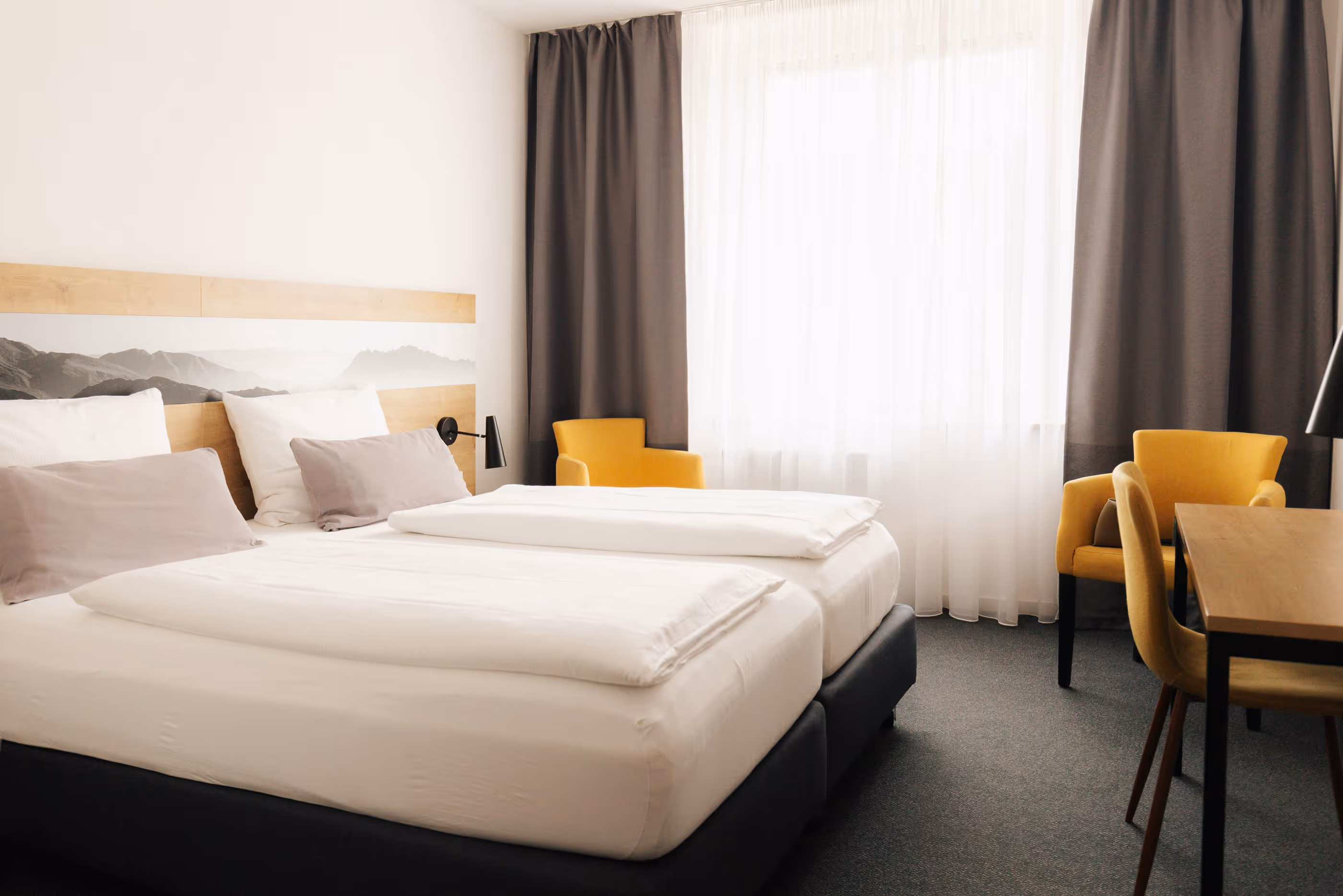 Hell eingerichtetes Hotelzimmer mit zwei Betten, gelben Sesseln, Schreibtisch und großen Fenstern mit weißen und grauen Vorhängen.