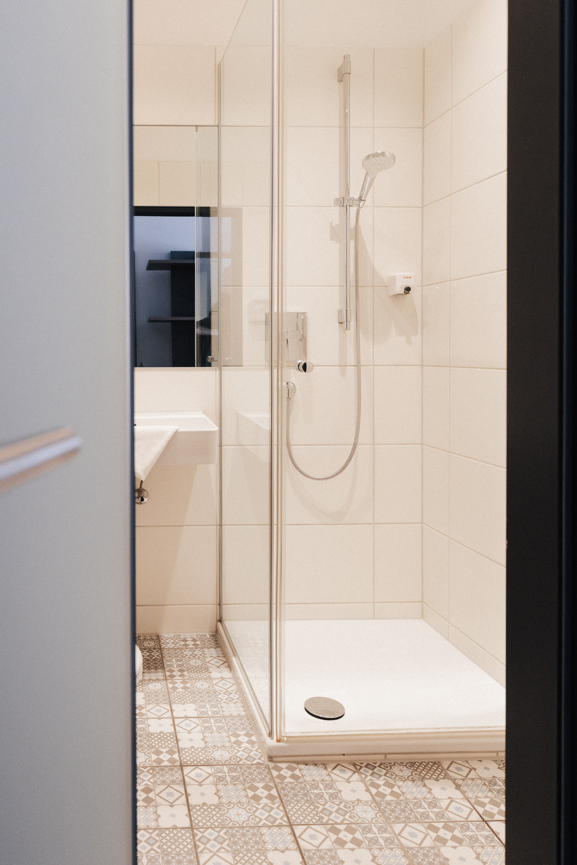 Helle Badezimmerdusche mit Glastür und gemustertem Fliesenboden.