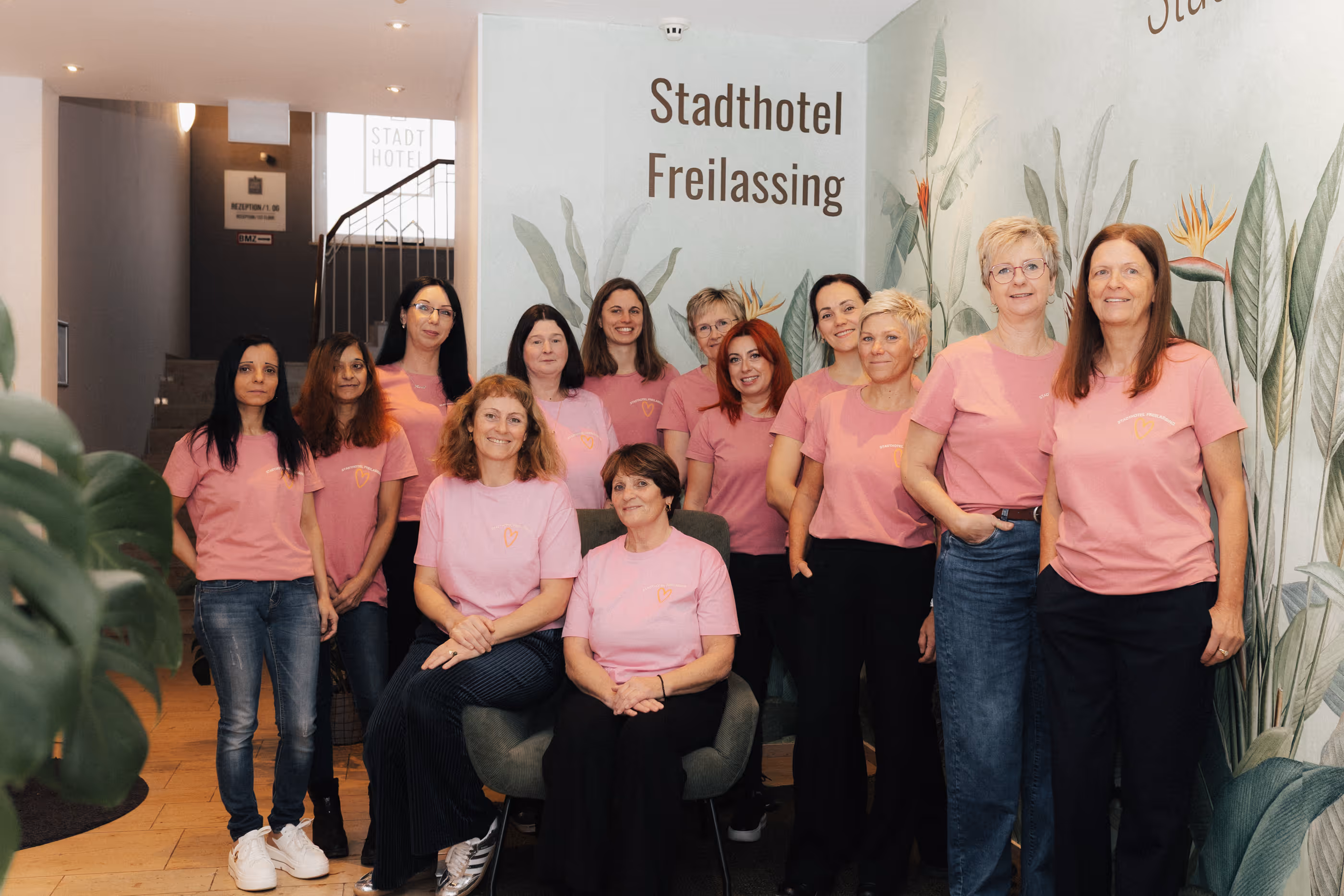 Gruppenfoto von zwölf Mitarbeiterinnen des Stadthotels Freilassing in rosa T-Shirts vor einer Wand mit botanischem Design.