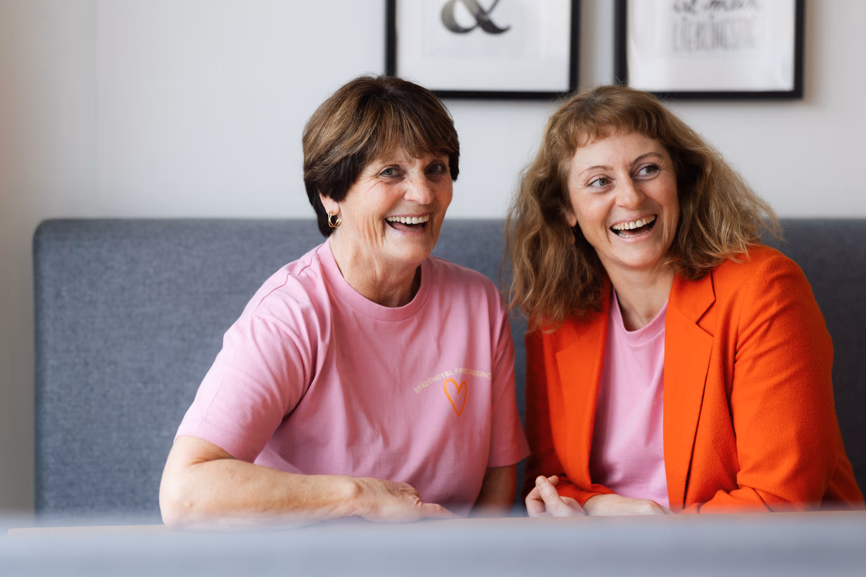 Zwei lächelnde Frauen sitzen nebeneinander auf einer grauen Bank, eine trägt ein pinkes T-Shirt, die andere ein pinkes T-Shirt und ein oranges Sakko.