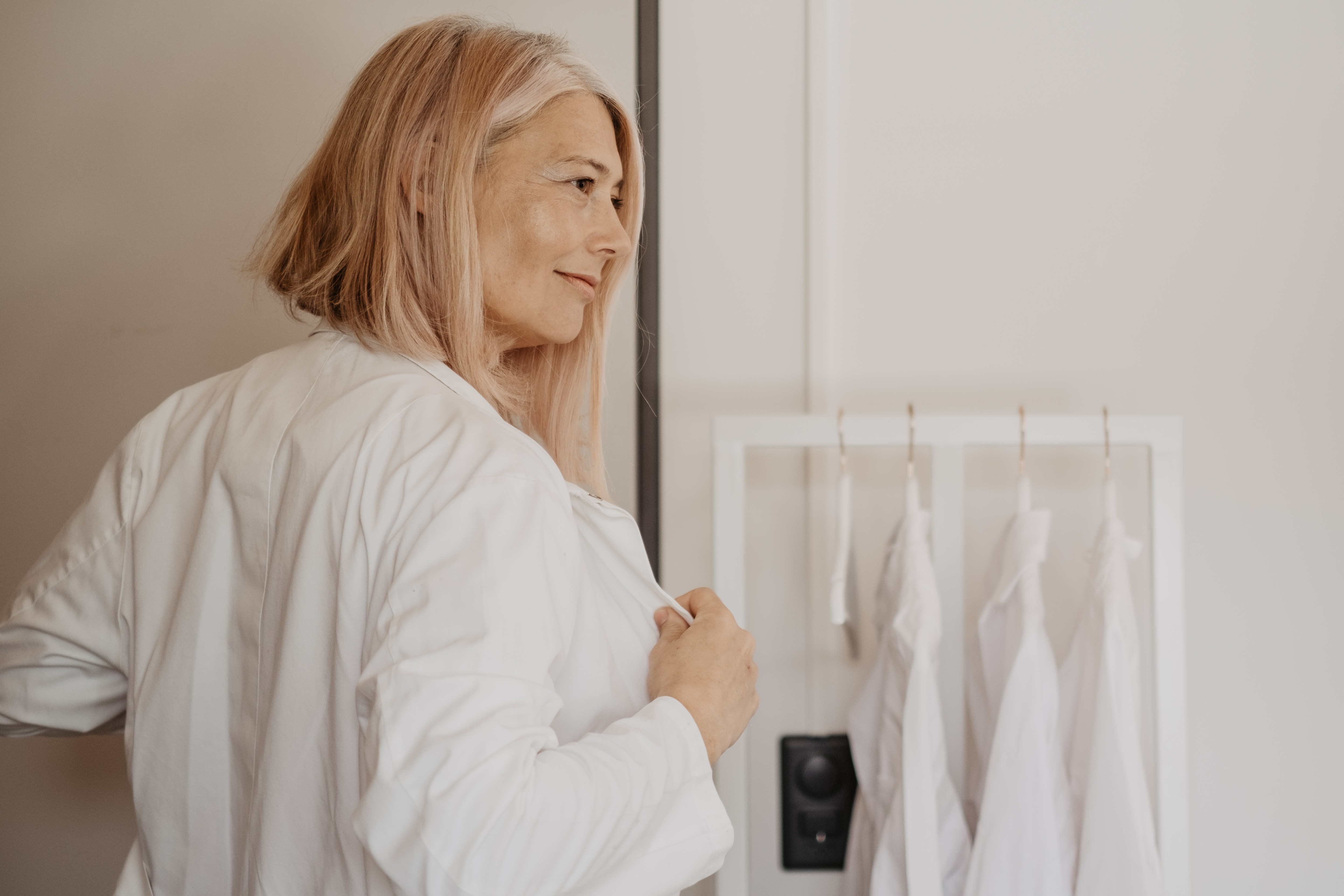 Judith Fiedler zieht sich gerade einen weißen Kittel über und steht vor der Garderobe. Sie schaut rechts aus dem Bild raus.