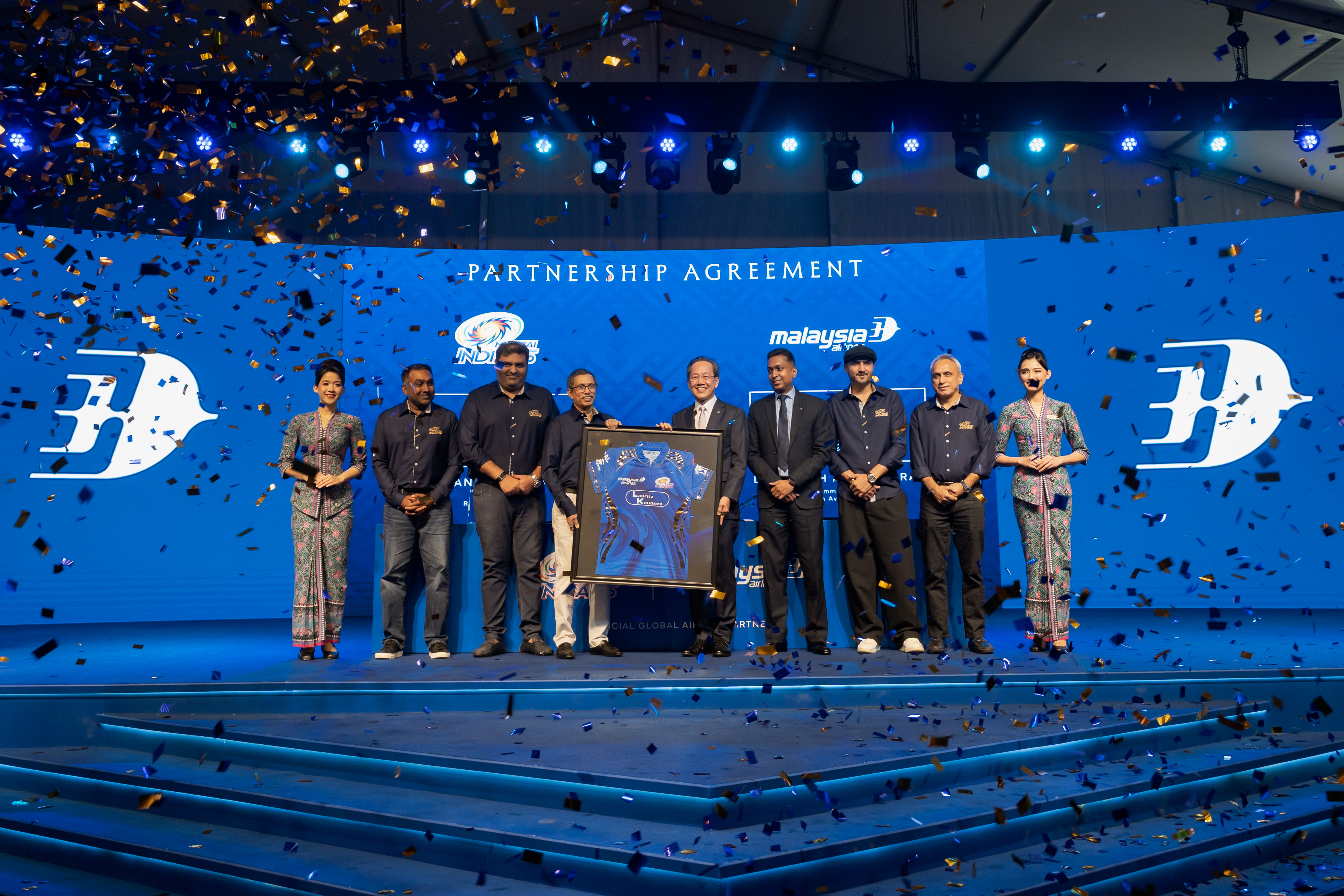 Group on stage holding framed blue sports jersey under confetti for partnership agreement event.