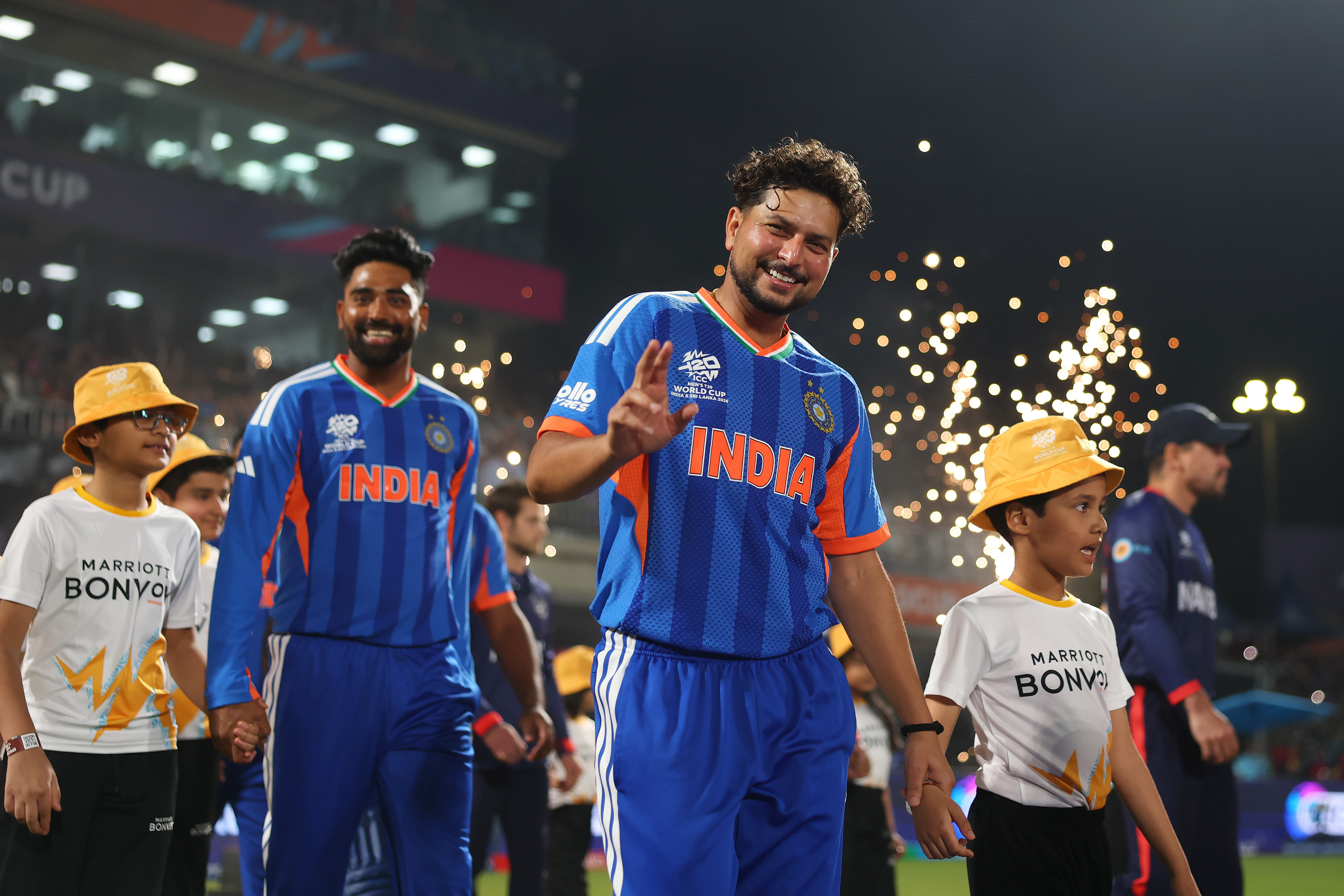 Indian cricket players in blue jerseys holding hands with children wearing Marriott Bonvoy shirts on field.