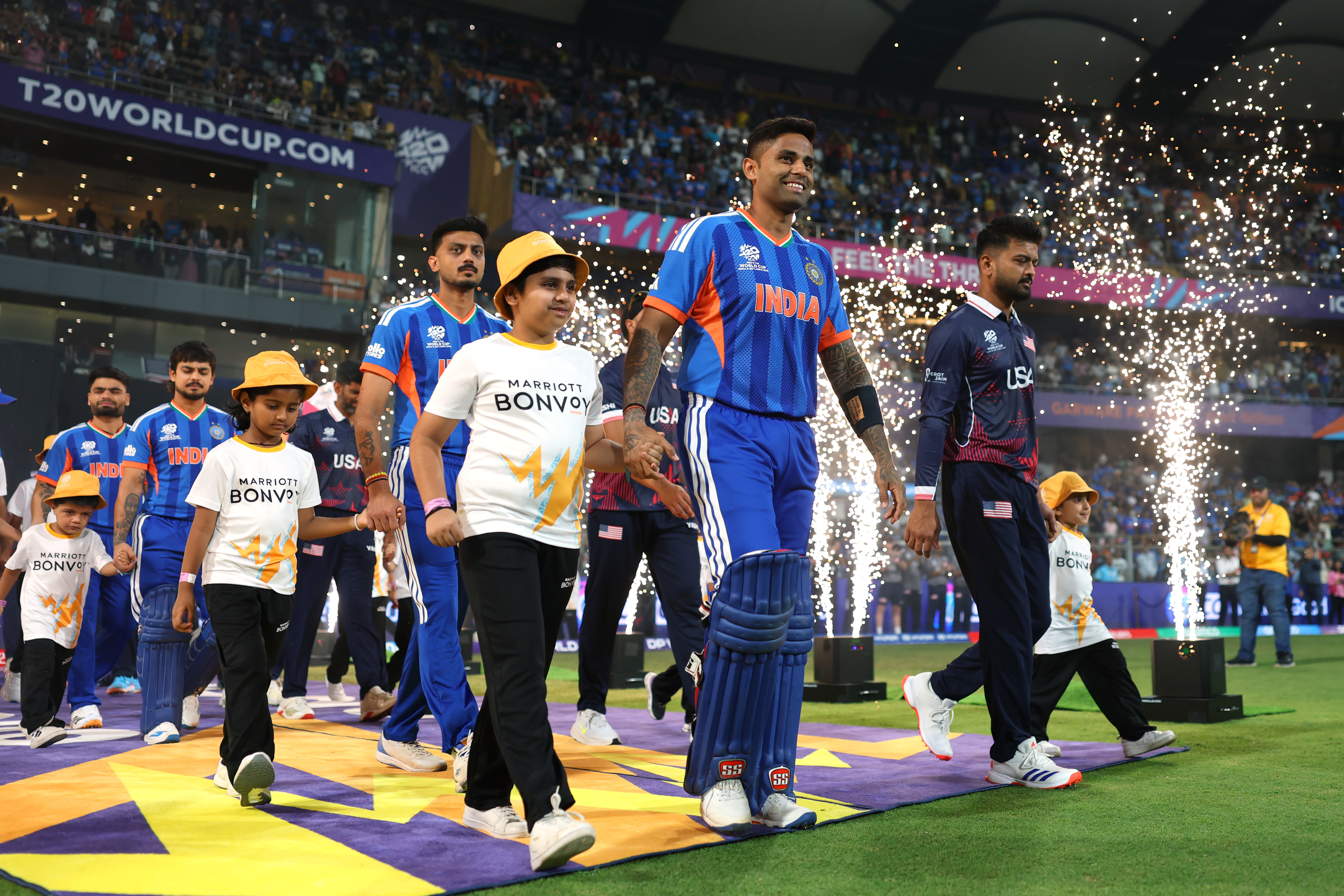 Indian and USA cricket teams walking on field with children as fireworks spark in the background.