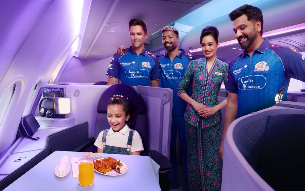 Smiling girl in airplane seat with meal and drink, three men in sports jerseys and a woman in uniform standing behind.