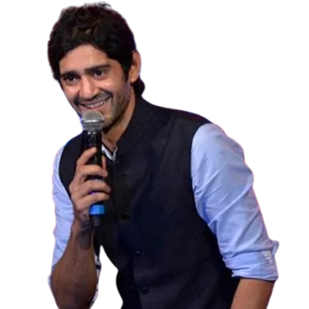 Smiling man with curly dark hair holds a microphone, wearing a dark vest over a light blue rolled-up shirt.