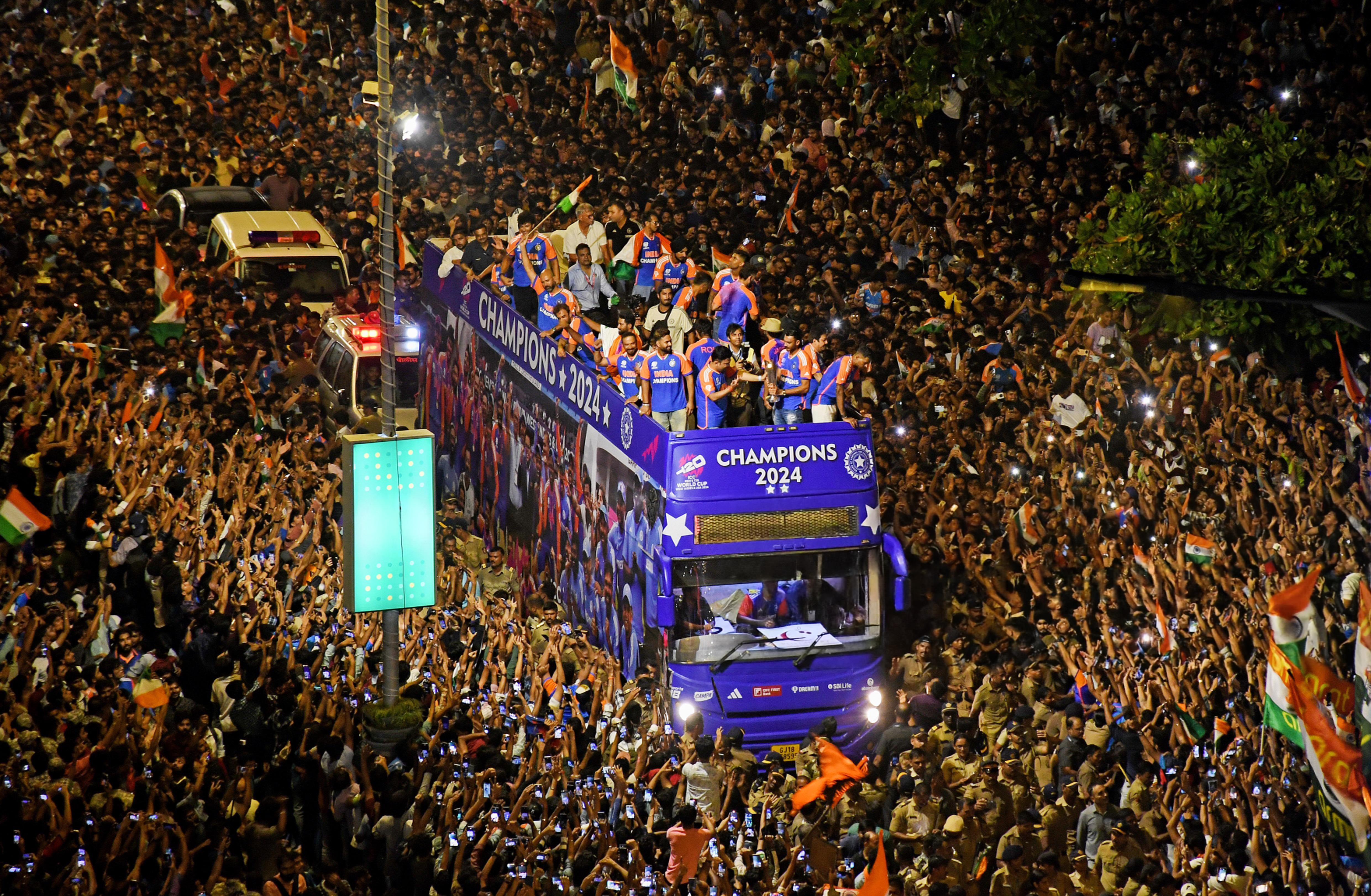 A large crowd celebrating around a purple double-decker bus displaying 'CHAMPIONS 2024' on the front and side, with people on top waving Indian flags in a nighttime parade.