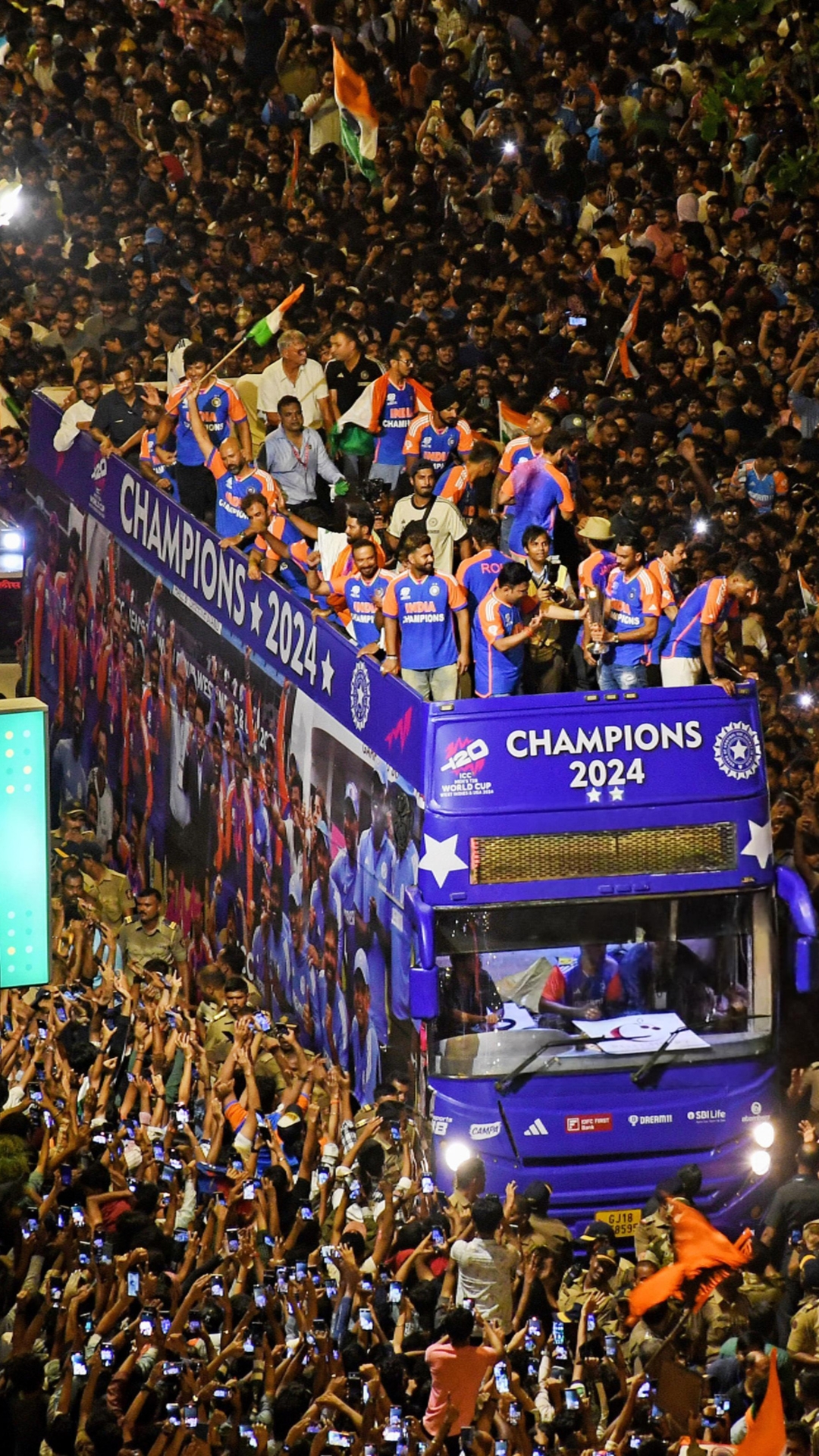 Celebratory parade with Indian cricket team members wearing blue and orange jerseys labeled 'INDIA CHAMPIONS' on a bus decorated with 'CHAMPIONS 2024' banners and logos for ICC T20 World Cup, surrounded by a large crowd of enthusiastic fans waving Indian flags and taking photos.