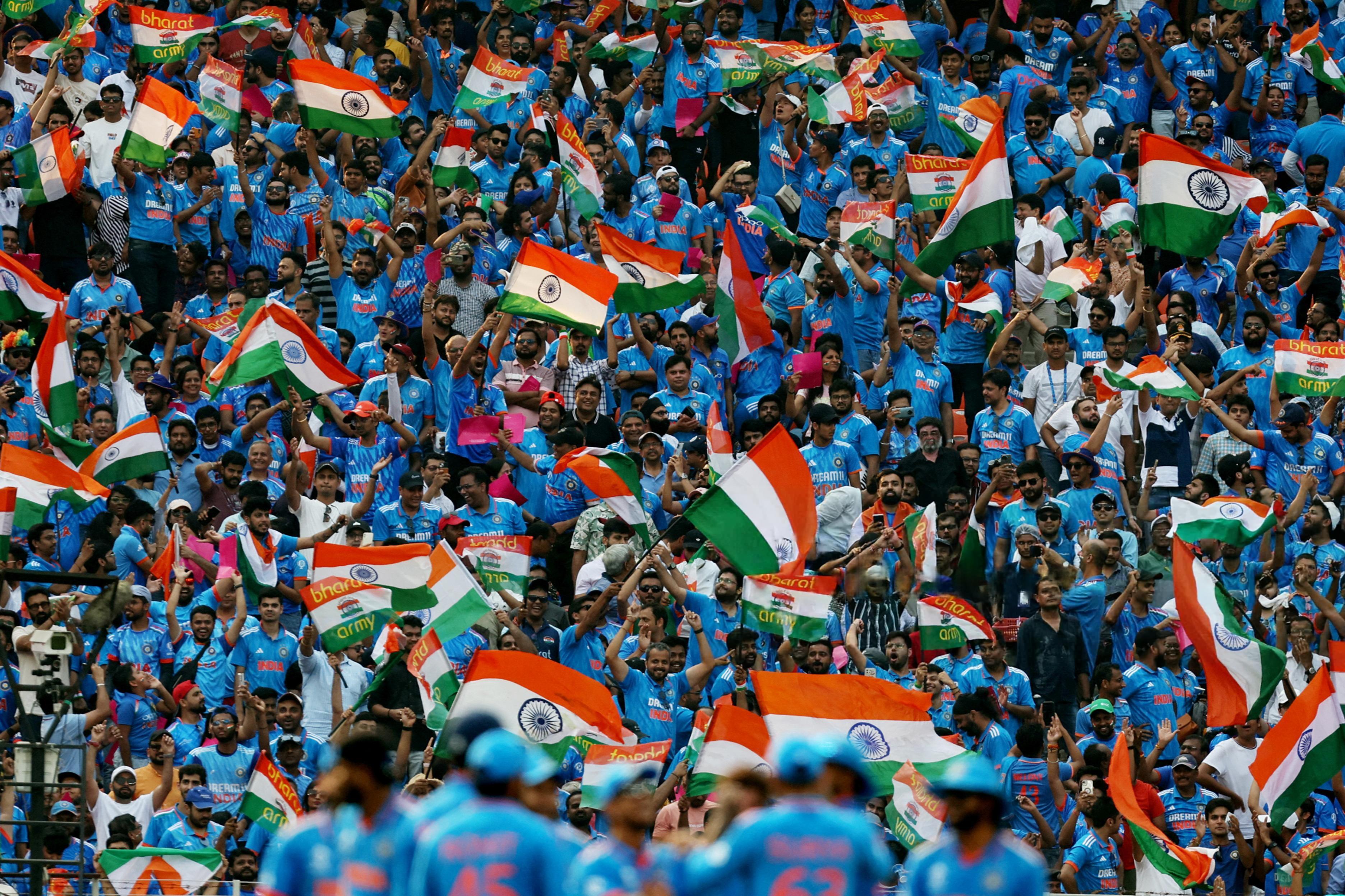 Large crowd of Indian cricket fans in blue jerseys enthusiastically waving numerous Indian national flags and banners with 'Bharat Army' text at a stadium, with some players blurred in the foreground.