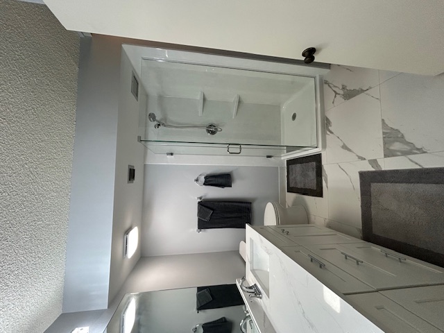 Modern bathroom with glass-enclosed shower, white vanity with sink, toilet, and dark gray towels and rugs.