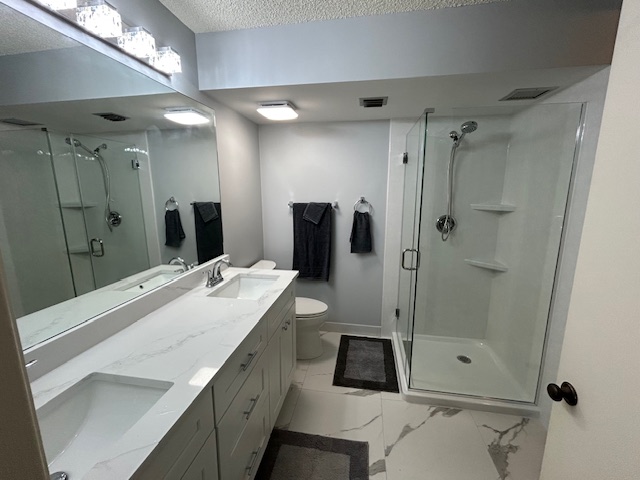 Modern bathroom with double sinks, large mirror, white countertop, glass-enclosed shower, toilet, and dark gray towels and rugs.