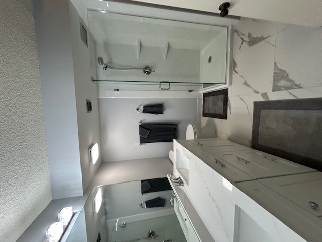 Modern bathroom with large mirror, white vanity with sink, glass shower enclosure, toilet, and gray towels and rugs.
