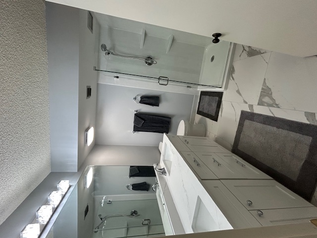 Modern bathroom with white cabinetry, double sink, large mirror, glass-enclosed shower, toilet, and gray bath mats and towels.