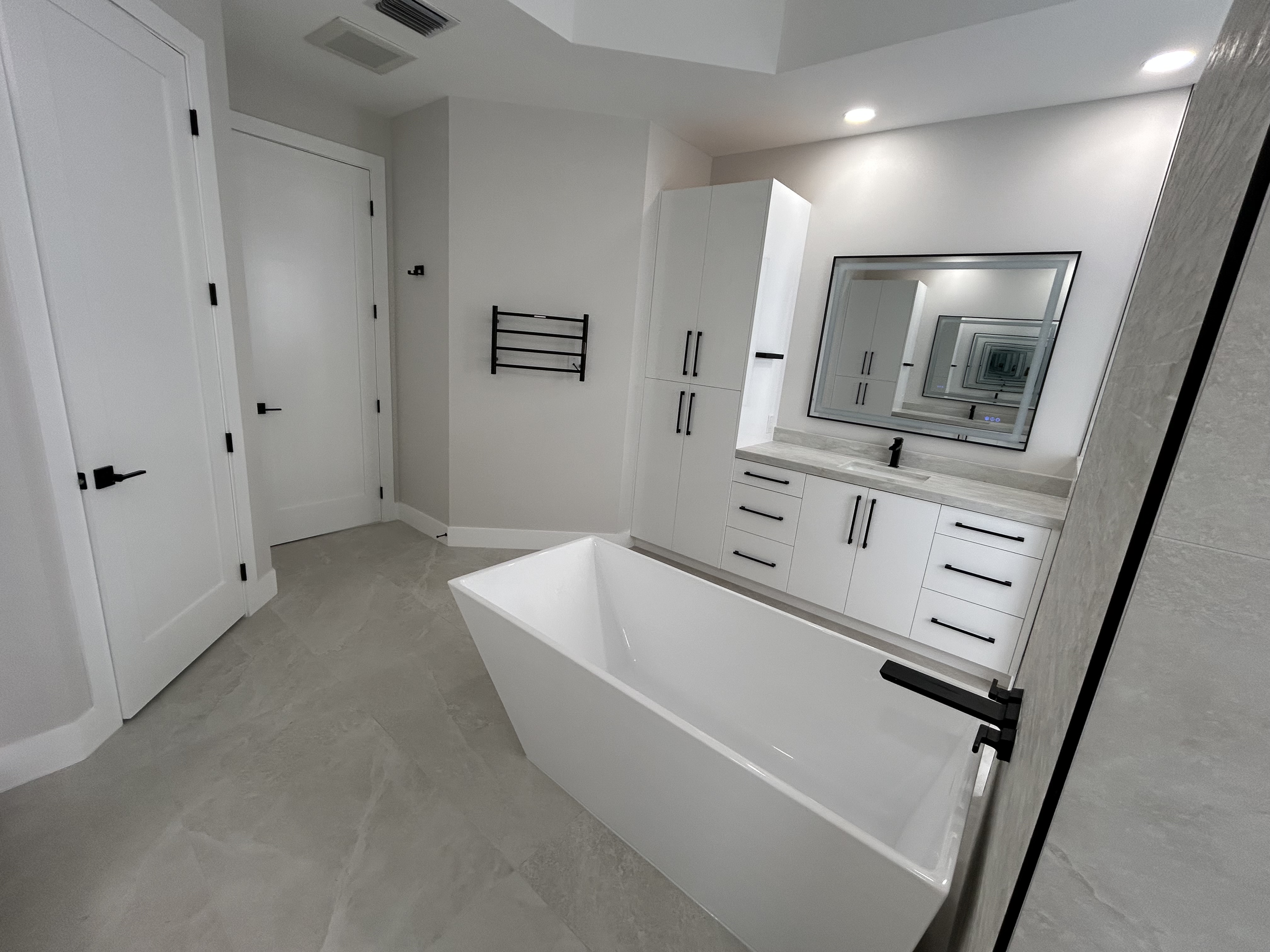 Modern bathroom with double sinks, large mirror, white countertop, glass-enclosed shower, toilet, and dark gray towels and rugs.