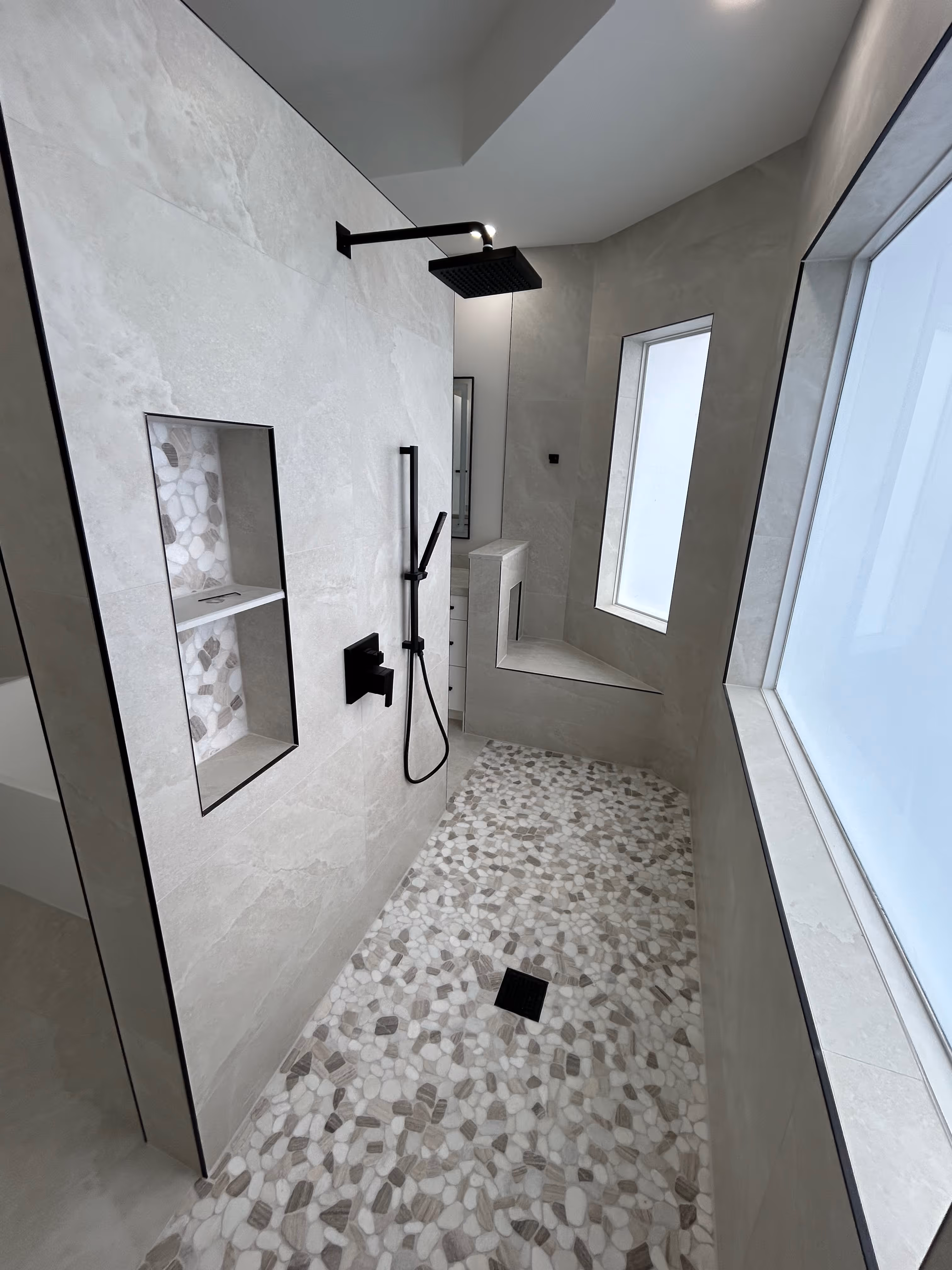 Modern bathroom with double sinks, large mirror, white countertop, glass-enclosed shower, toilet, and dark gray towels and rugs.