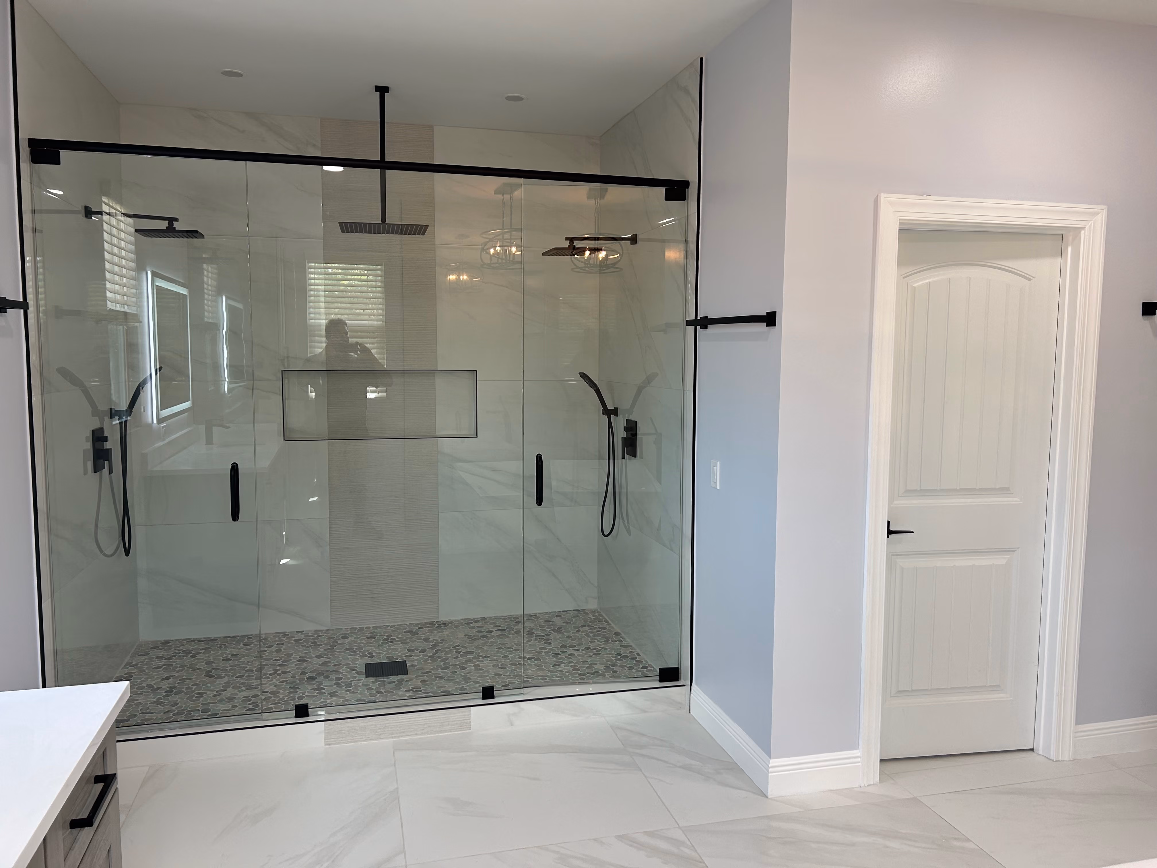 Modern bathroom with large glass shower enclosure featuring two black rain showerheads and pebble stone floor.