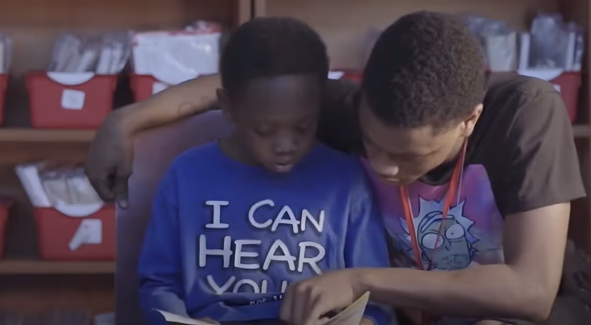 Two black boys, on of them a little older mentoring a younger one, teaching him how to read. He has his shoulder over him.
