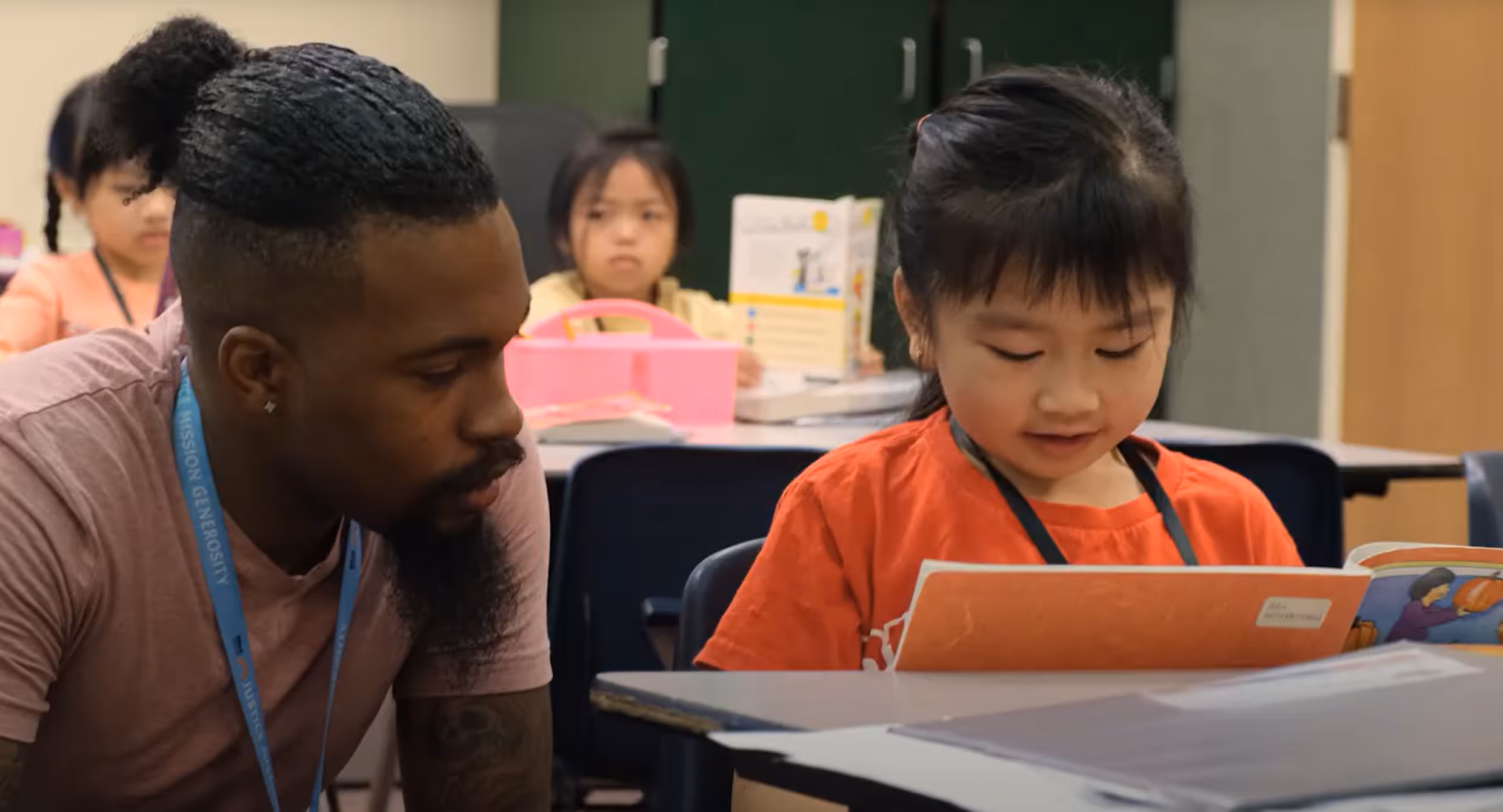 A mentor from a faith-based non-profit helps a young student read in a classroom, supporting literacy development and connection through education.